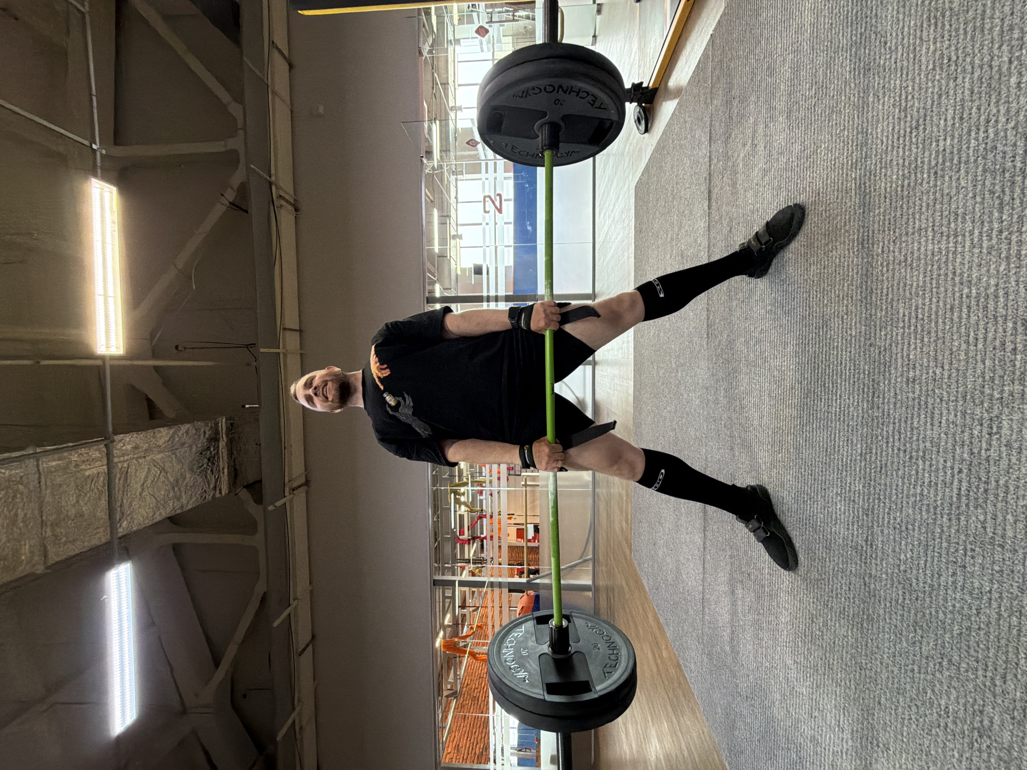 A person is standing in what appears to be a gym in the city of Chaikovsky, Russia. The individual is wearing a black t-shirt, black shorts, black knee-high socks, and black shoes. They are holding a barbell loaded with weights and are smiling. The gym has a light-colored carpet, and there's a window view showing orange roofing and other building structures, typical of Chaikovsky's urban environment. Overhead, there is a visible industrial-style ceiling with ductwork and a strip light. The weights on the barbell have the brand "TECHNOGYM" visible on them. The person has straps wrapped around their wrists connecting them to the barbell.