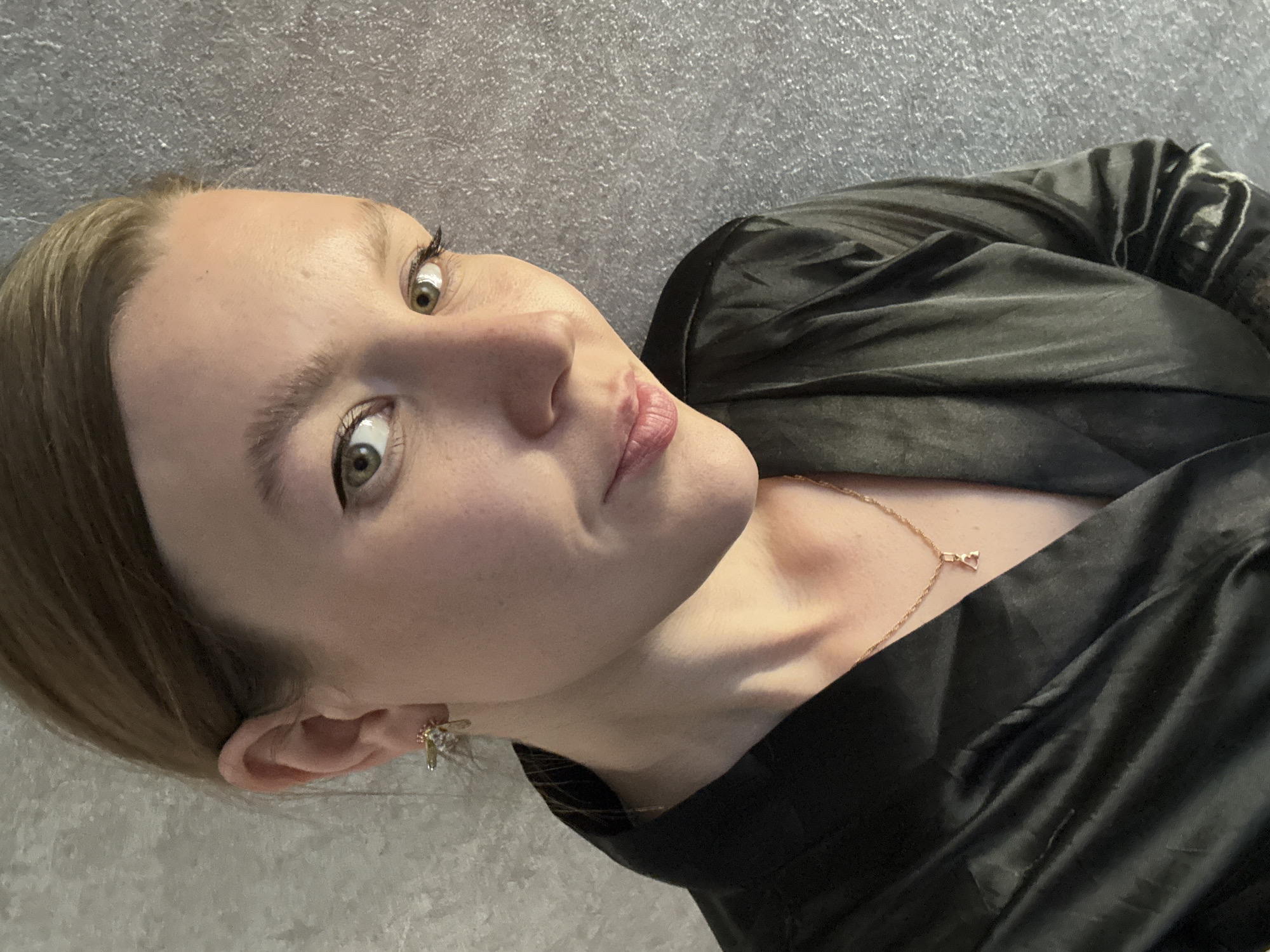 A woman with fair skin, light brown hair, and green eyes is featured in a selfie-style photograph. She has a subtle expression on her face with slightly parted lips. She is wearing a black, silky garment that appears to be a robe or jacket. A thin gold chain necklace with a heart-shaped pendant is visible on her neck. She also has a pair of small, floral-shaped earrings. The background is a textured gray surface, potentially a wall. There is no indication of the location being Izhevsk, Russia, or any other specific location.