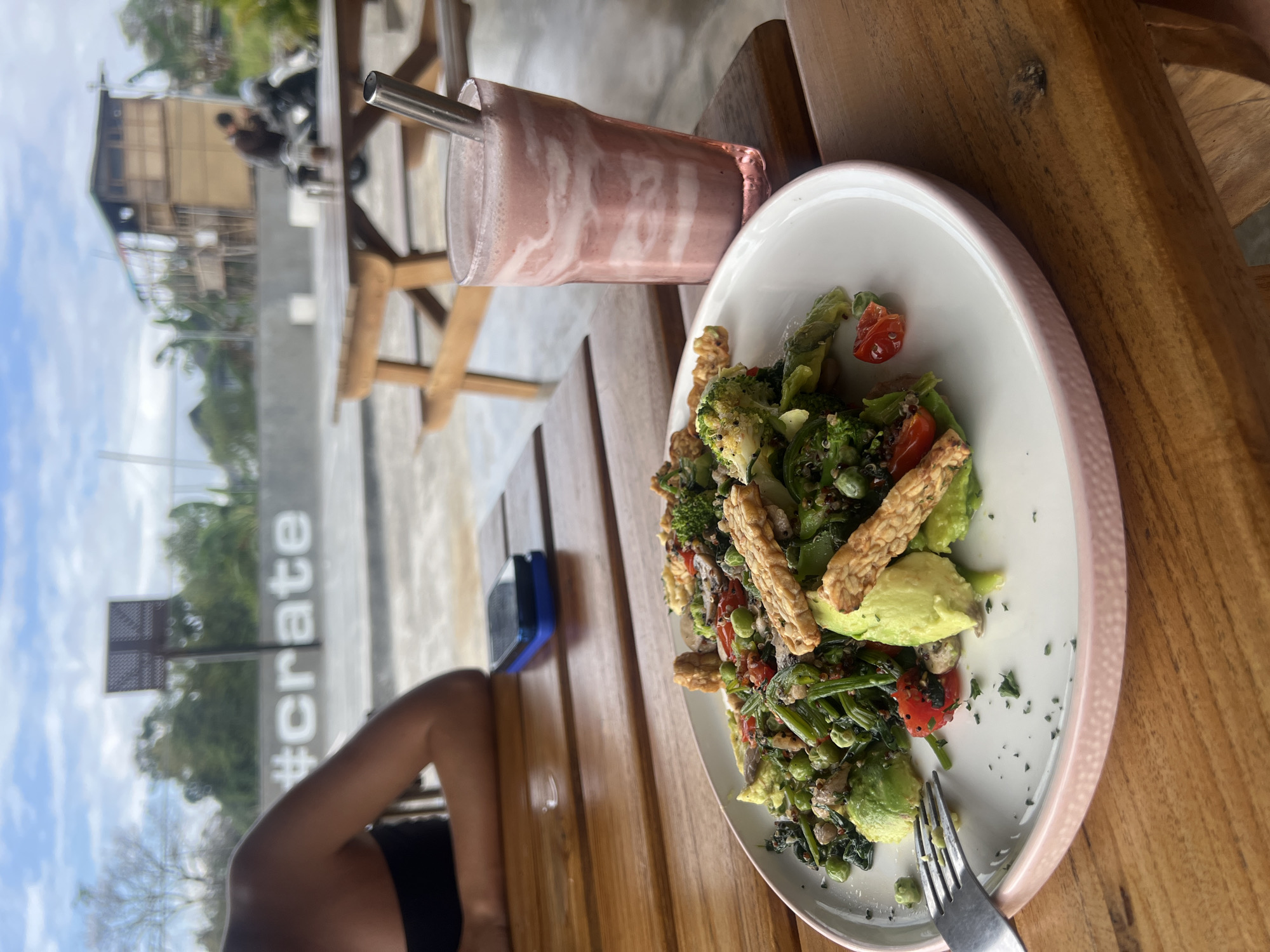 A close-up, slightly tilted shot shows a healthy meal and drink on a wooden table. In the foreground, a white plate with a pale pink rim holds a vibrant salad. The salad is a mix of green vegetables, including broccoli florets, edamame, and leafy greens. Slices of avocado and pieces of what appear to be tempeh are also prominent. A few cherry tomatoes are scattered throughout. A fork rests on the edge of the plate, with some stray herbs sprinkled on the white surface.
To the left of the salad, a clear glass contains a pink, creamy smoothie, swirling with lighter pink or white streaks. A silver metal straw extends from the glass. The drink appears to be a blended fruit beverage.
The background is blurred but provides context. A wooden slatted table and chairs are visible, suggesting an outdoor seating area. Beyond that, a building with a prominent sign is visible, with the text "#crate" clearly legible. The sky is cloudy, indicating daylight. The overall impression is a relaxed, healthy meal in a casual setting, likely a cafe or restaurant, given the sign. The image appears to have been taken during the day, with natural light.