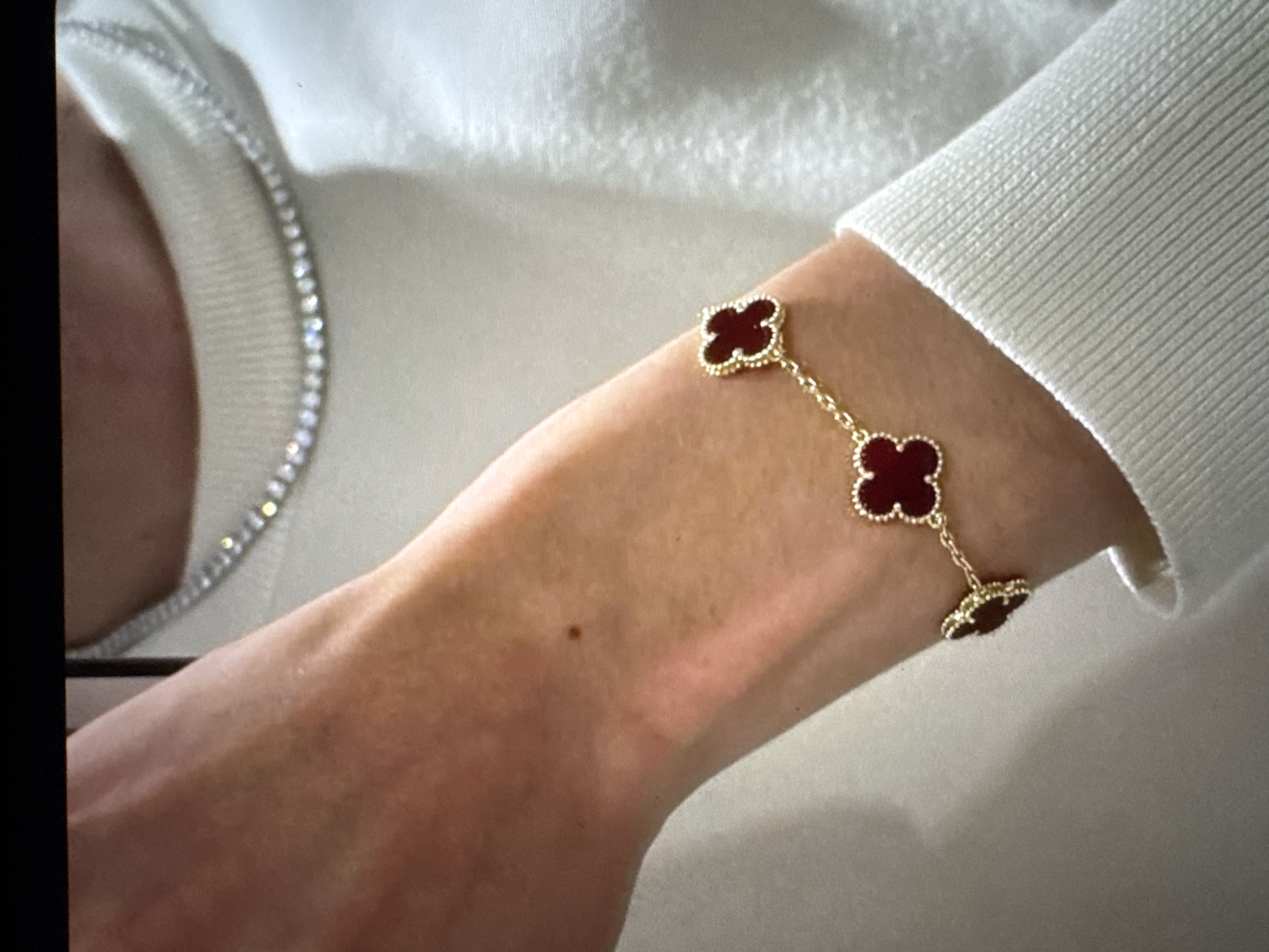 A close-up shot focuses on a person's arm wearing a gold bracelet with red clover-shaped charms. The bracelet is positioned on the forearm, with three charms visible. The person is also wearing a white crew-neck sweater and a thin, sparkling necklace. The background is out of focus and appears to be indoors, with soft lighting. No details about the city of Tallinn, Estonia, are present in the image.