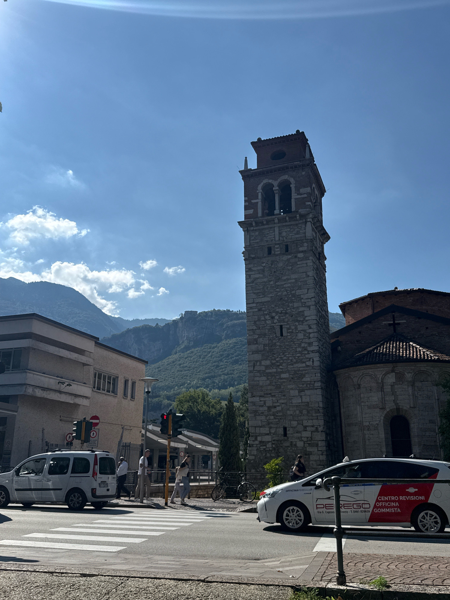 The image captures a street scene in Trento, Italy, dominated by a tall, stone bell tower and a portion of an ancient church. The bell tower is constructed from large, roughly hewn stone blocks and features arched openings near its top, topped by a tiled roof. Adjacent to it, the church exhibits a rounded apse with a tiled roof and a stone facade. In the foreground, a white passenger van is parked near a crosswalk, and a white car with red and blue decals, identified as "CENTRO REVISIONI OFFICINA GOMMISTA" with "PEREGO" prominently displayed, is driving past. Several pedestrians are visible on the sidewalk, and a traffic light displays a red signal. The background reveals lush green mountains under a bright blue sky with scattered white clouds, suggesting a clear, sunny day. The architecture and the surrounding landscape are characteristic of Trento.