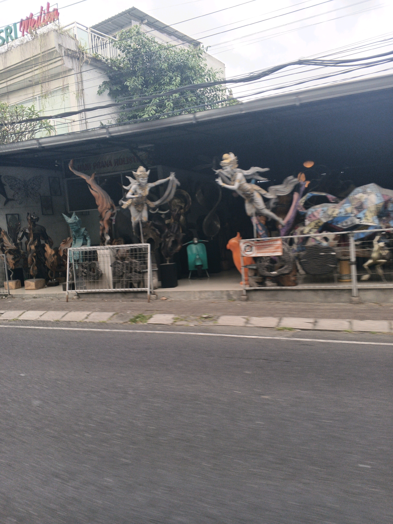 The image shows a storefront in Ubud District, Indonesia, with a variety of sculptures and decorations displayed. The storefront appears to be a shop selling art or souvenirs, with a large, colorful sculpture prominently featured. The background shows a building with a sign that reads "SRI Melatih." The scene is outdoors, with a street in the foreground. The overall impression is one of a bustling commercial area.
