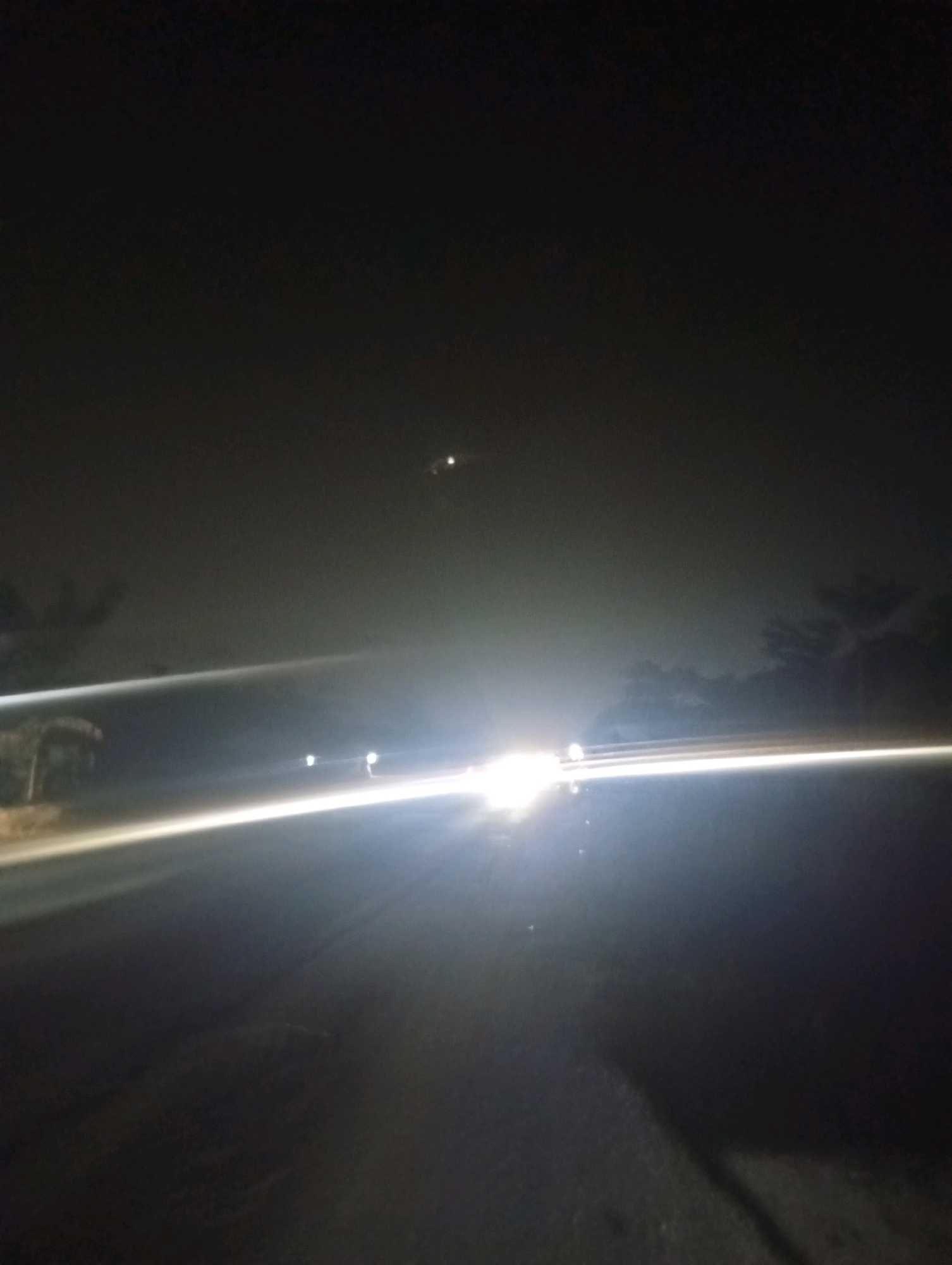 The image depicts a nighttime scene on a road, possibly in or near Kubang Jaya, Indonesia. The foreground is a dark, asphalt road. A bright light streaks horizontally across the mid-ground, suggesting the motion blur of headlights from a vehicle. This bright streak obscures much of the road and the environment. In the distance, there are faint silhouettes of trees and what appear to be other lights, possibly from streetlights or other vehicles. The sky is dark and hazy, with a small, bright, circular object visible high in the frame, which could be a distant light source or a celestial body. The overall impression is one of low visibility and movement on a road at night.
