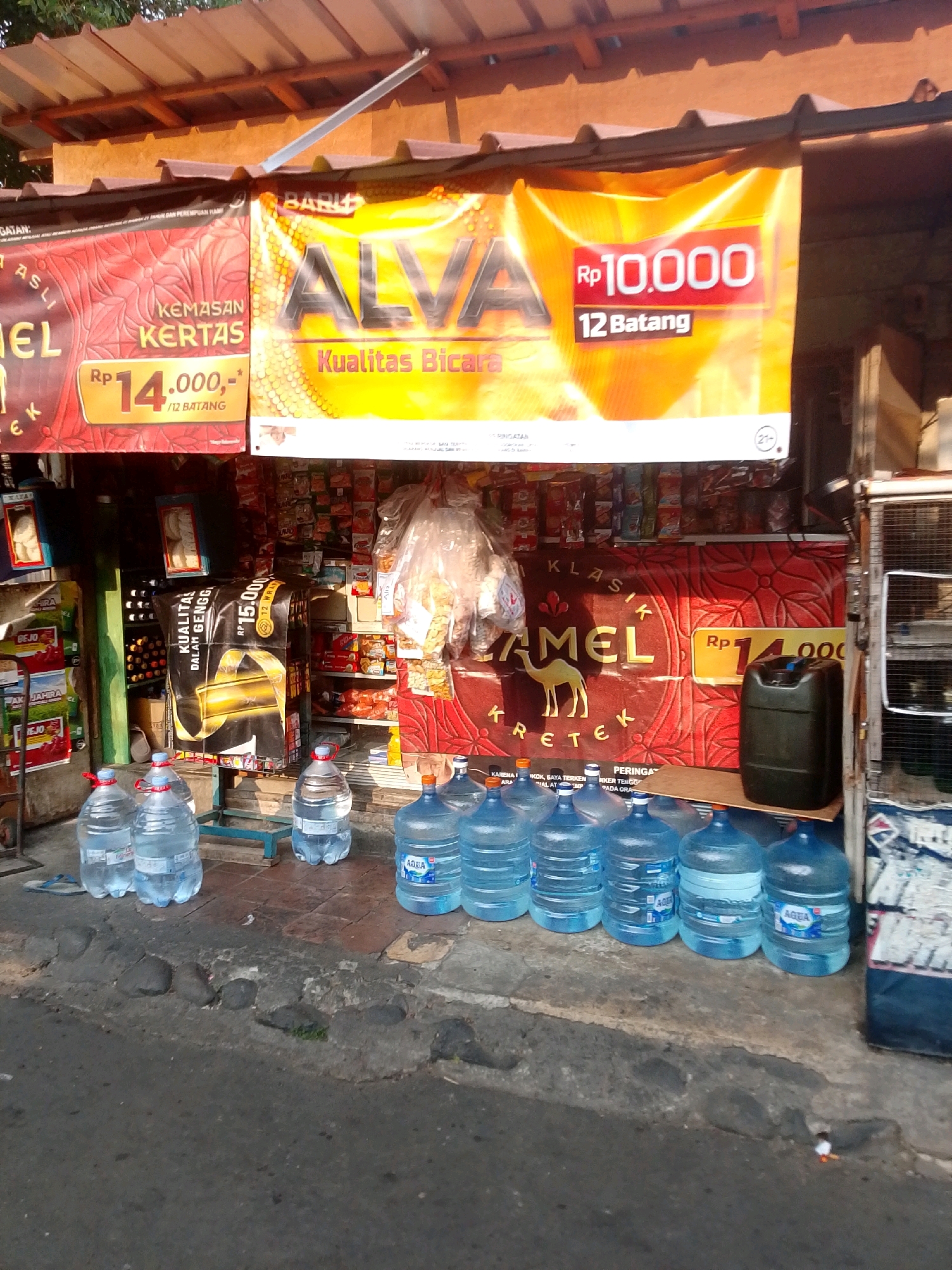 The image depicts an outdoor street-level view of a small retail stall in East Jakarta, Indonesia, likely a "warung" or convenience store. The stall is adorned with prominent advertising banners, one in yellow featuring "ALVA" and another in red with a "CAMEL" brand logo, both indicating cigarette promotions with prices displayed in Indonesian Rupiah. In front of the stall, a line of large, clear plastic water jugs, some with "AQUA" branding, are arranged on the ground. Behind the water jugs, various goods are displayed on shelves inside the stall, including packaged snacks and beverages. The ground in front of the stall is paved with a combination of concrete and dark stones. The overall scene suggests a typical small business operating in a local neighborhood.