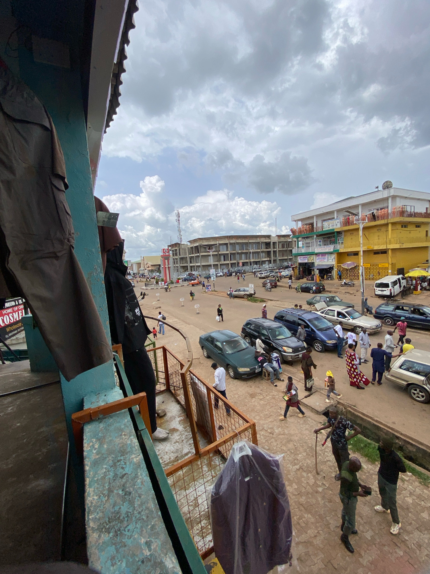 This is a high-angle, medium shot of a bustling street scene in Jos, Nigeria, likely near the central market or a commercial hub. The foreground features a balcony or overhead structure with a partial view of brown pants hanging on display and a black mannequin or garment. The street below is filled with various cars, including sedans and SUVs, parked and in motion. Numerous people are walking, interacting, and congregating on the sidewalks and street. Buildings line the street, with a prominent yellow building with multiple stories and balconies visible. The sky is overcast with dramatic clouds, suggesting a potentially rainy or humid day. The overall impression is one of vibrant urban activity.