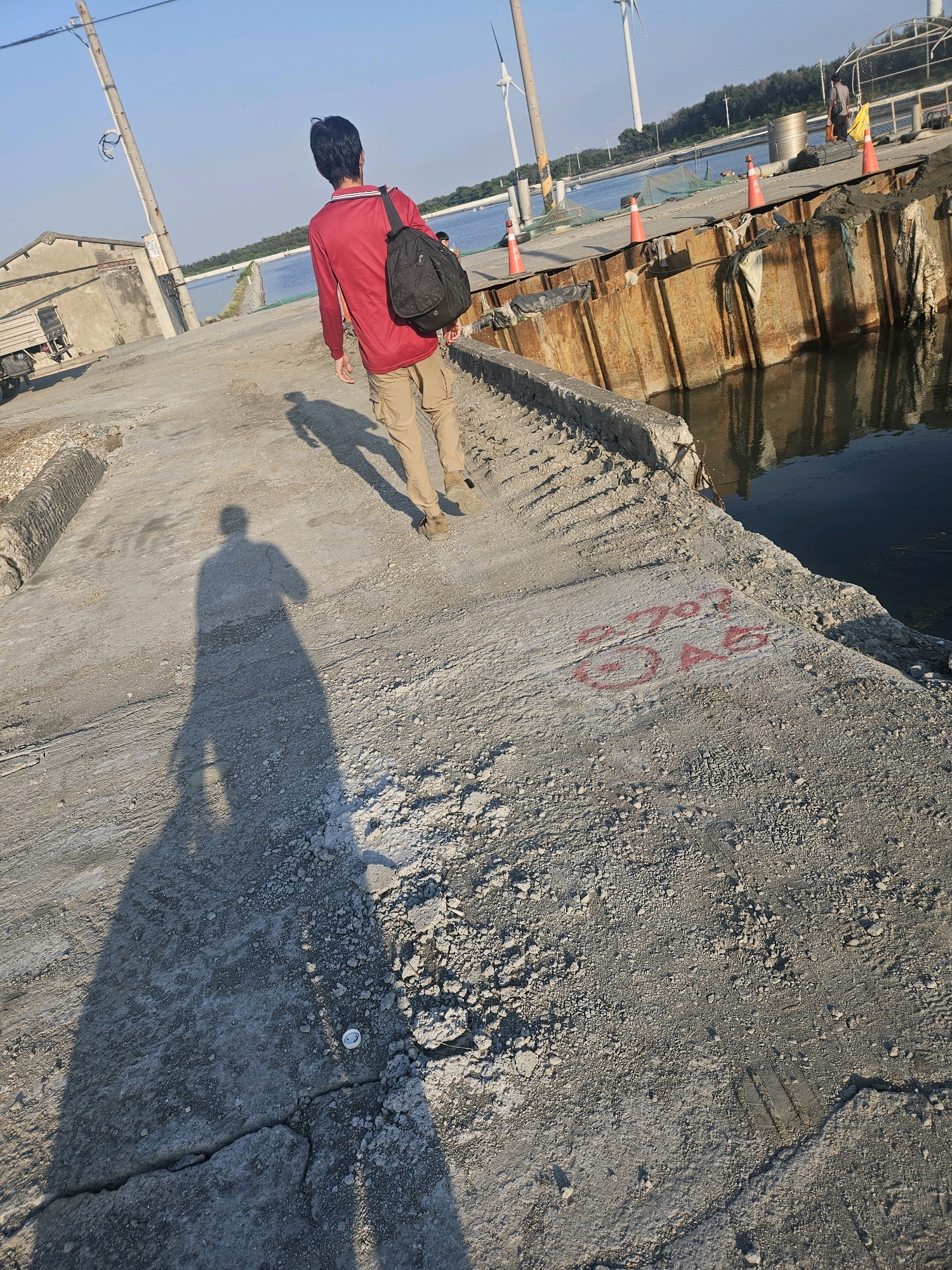 A person wearing a red long-sleeved shirt and khaki cargo pants, carrying a black backpack, walks away from the camera on a concrete path. The path leads to a body of water with a rusty metal seawall. In the background, there are several wind turbines, some buildings, and a clear blue sky. The scene appears to be in Taixi Township, Taiwan. The sun casts a long shadow of the person on the concrete. There are red markings on the concrete in the lower right portion of the image.
