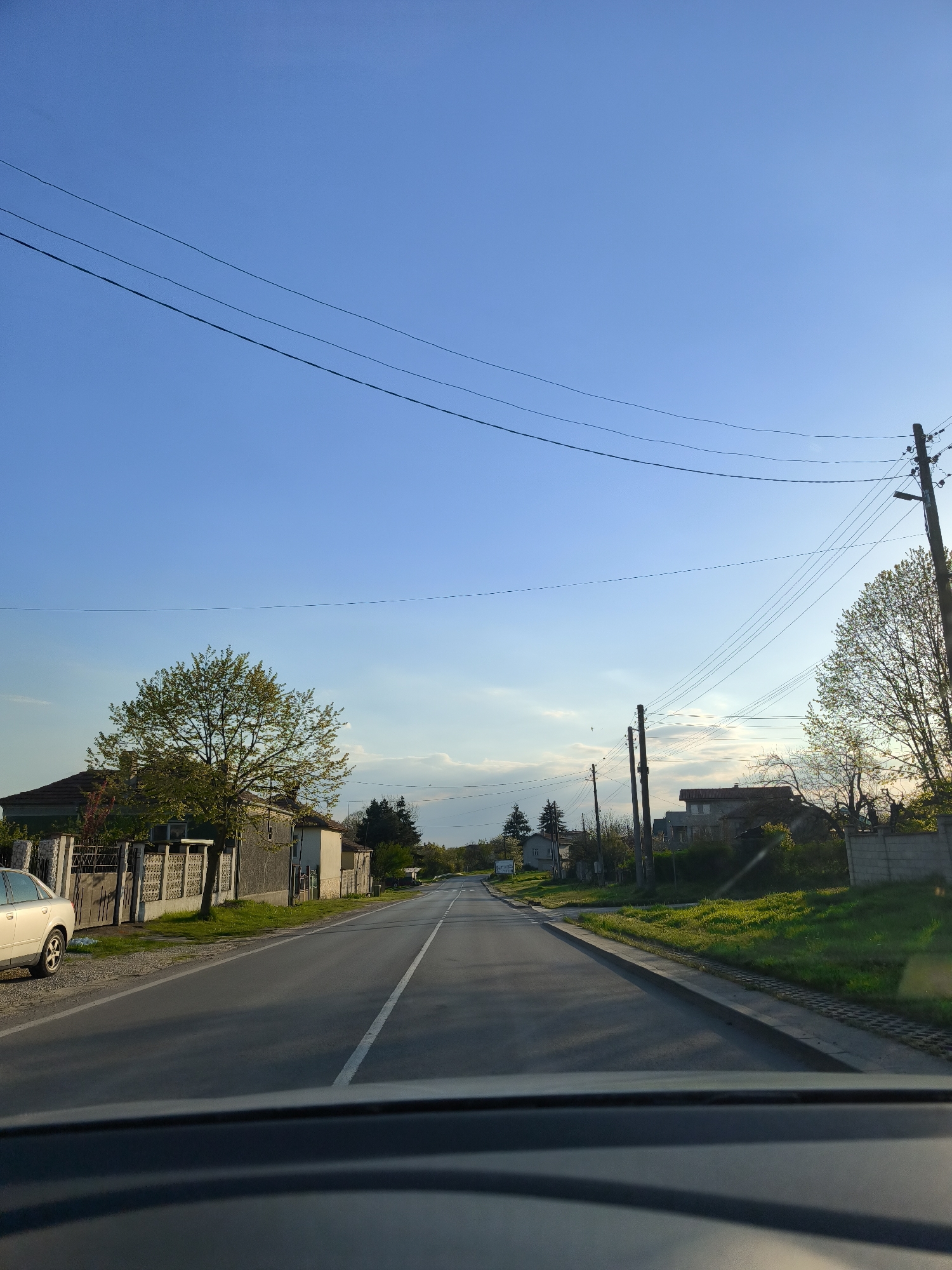 Captured from within a vehicle, the photograph depicts a residential street in Zdravets, Bulgaria. The asphalt road is characterized by a single white lane marker bisecting its width. On the left, a parked silver-colored car faces forward, next to fenced houses with trees in the yard. Electrical poles and wires traverse the sky above. The sky is blue. The scene is captured in daylight, but the lighting is beginning to indicate the oncoming evening. The image is taken from the driver's position.