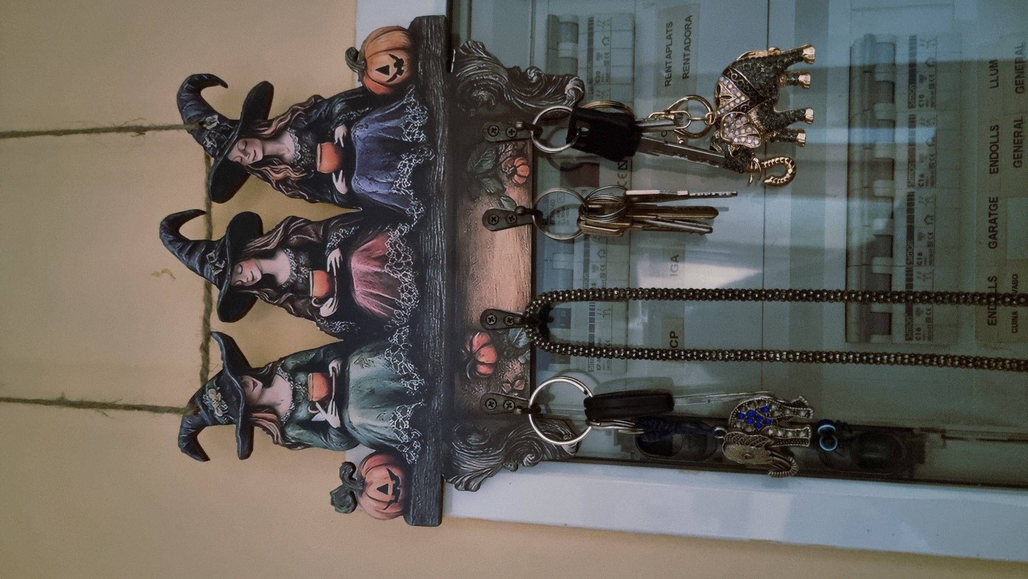 In Sant Joan Les Fonts, Spain, a decorative wall-mounted key holder is shown. The key holder features three stylized figures of witches in black pointed hats, each holding a mug. They are seated on a dark-colored base adorned with small pumpkin decorations. Below the witch figures, there are three hooks. The left hook holds a keyring with two keys and a blue-jeweled elephant charm. The middle hook has a long, dark beaded chain. The right hook supports a keyring with several keys and a gold-colored elephant charm. The background consists of a light-colored wall and a white electrical panel with various switches and labels, one of which reads "RENTAPLATS RENTADORA." The lighting is ambient, casting soft shadows.