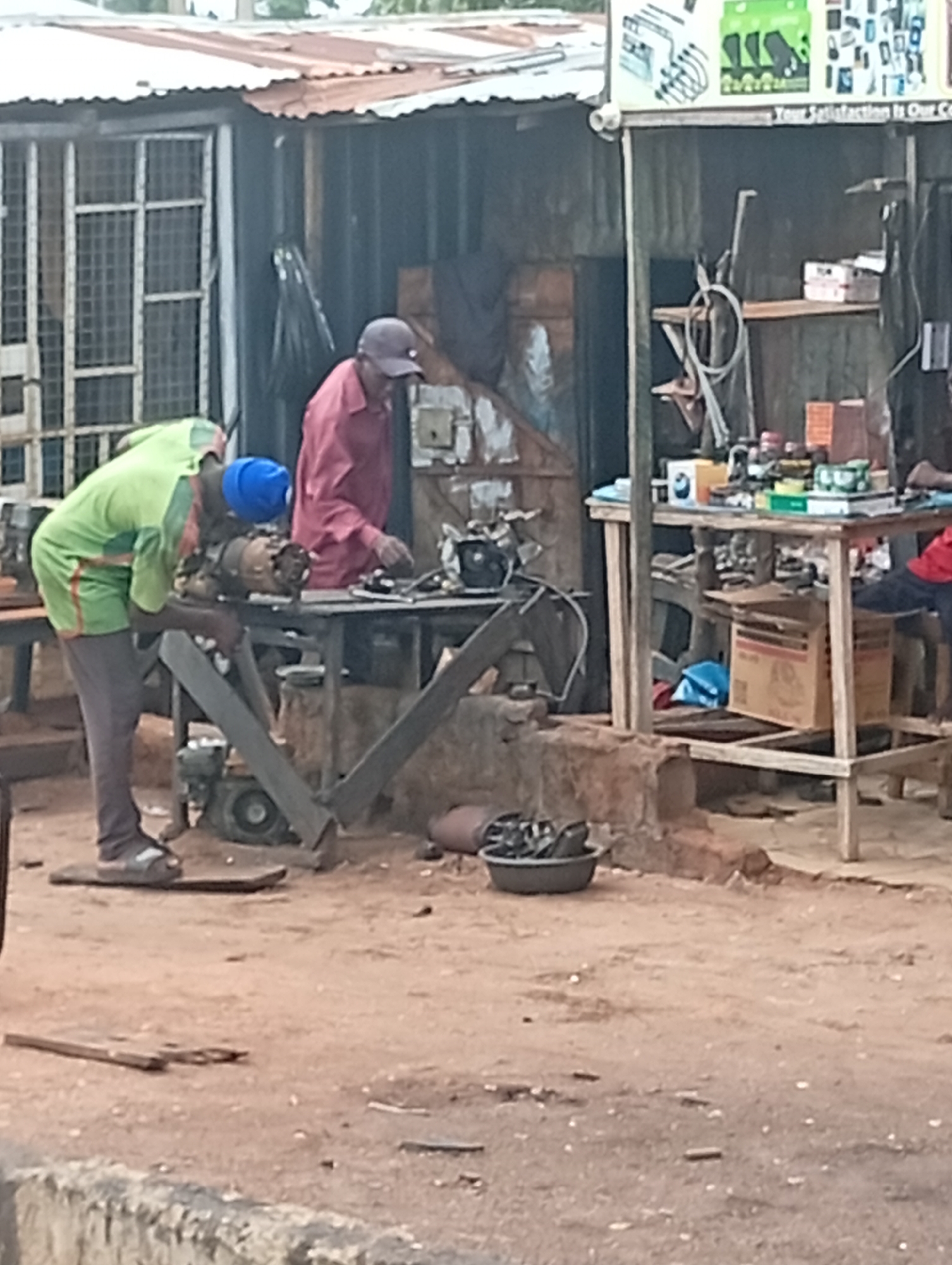 The image shows an outdoor workshop in Tumfure, Nigeria. Two men are present. The man on the left, wearing a green shirt and dark pants, is bent over a piece of machinery, appearing to be working on it. He has a blue cap on. The man on the right, wearing a pink shirt and dark pants, is standing behind a table and also appears to be engaged in some activity. The workshop is cluttered with various objects, including tools, parts, and boxes. A corrugated metal structure forms the background, with shelves and more items stored within. The ground is dusty earth.