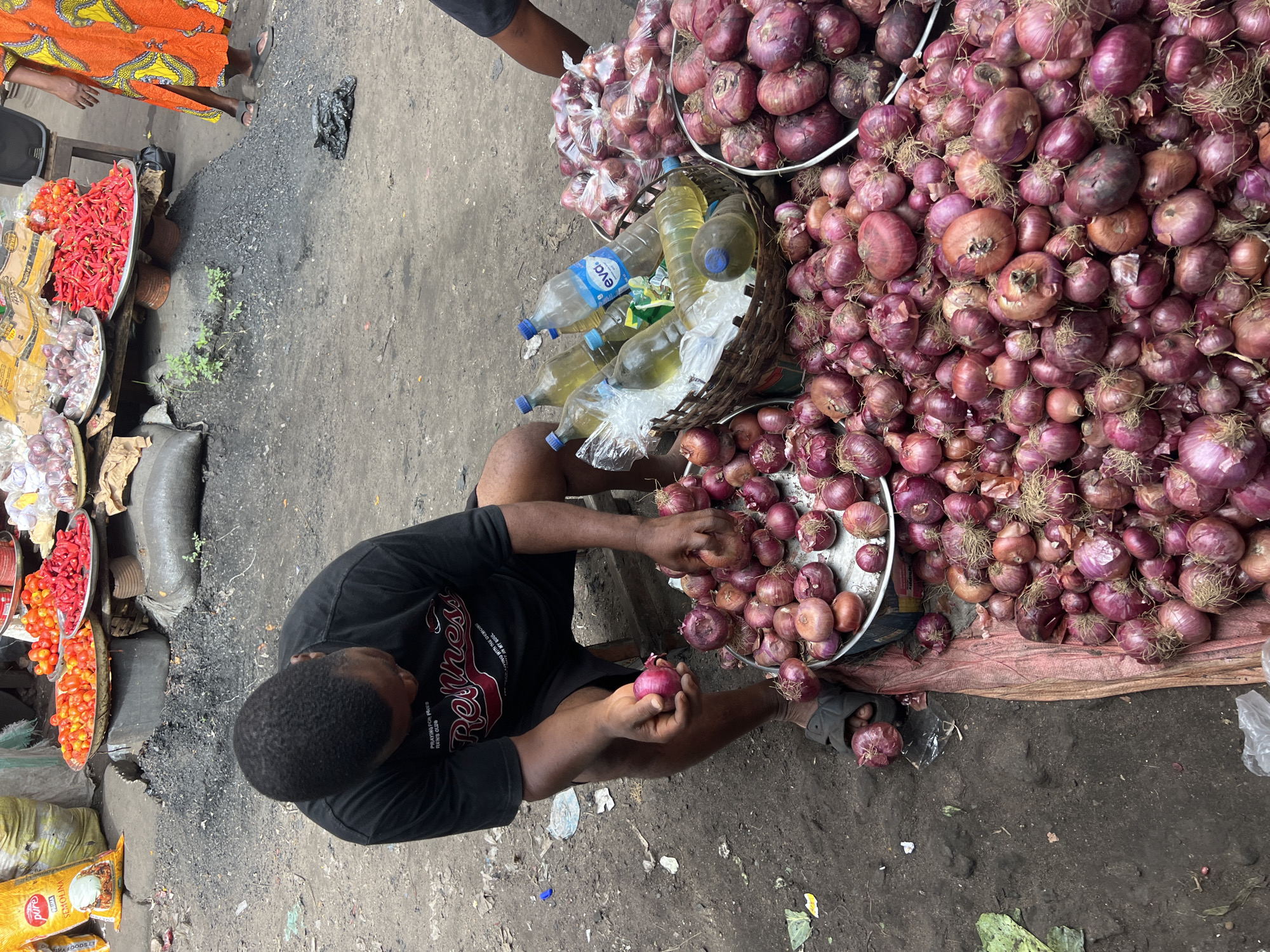 This image depicts a bustling outdoor market scene in Nkpor, Nigeria, showcasing a variety of fresh produce. A man, wearing a black t-shirt and dark shorts, is seated on the ground, engaged in sorting or preparing numerous red onions. He holds one onion in his hands, surrounded by large quantities of onions displayed in metal trays and directly piled on a reddish-brown tarp.
Adjacent to him, other market offerings are visible, including metal trays overflowing with bright red chili peppers and smaller red tomatoes. Also present are garlic bulbs, pre-packaged onions in clear plastic bags, and a prominent yellow bag labeled "PRIMA SEMOLINA." A wicker basket holds several plastic bottles, some containing clear liquid and others a golden cooking oil, with at least one bottle bearing the "eva." label.
The ground surface is a mix of dirt and concrete, characteristic of a busy market area, with scattered debris such as charcoal, plastic, and leaves. In the upper left, the lower body of another person, clad in an orange and yellow patterned garment and sandals, is partially visible. The scene is brightly lit, indicating daytime market activity.
Visible text elements include "Refresh," "PRAYING FOR P.C. NEWS CLUB," and "LIVING WITH THE DEMONS IN MY SOUL" printed on the man's t-shirt, "PRIMA SEMOLINA" on the yellow bag, and "eva." on the water bottles.