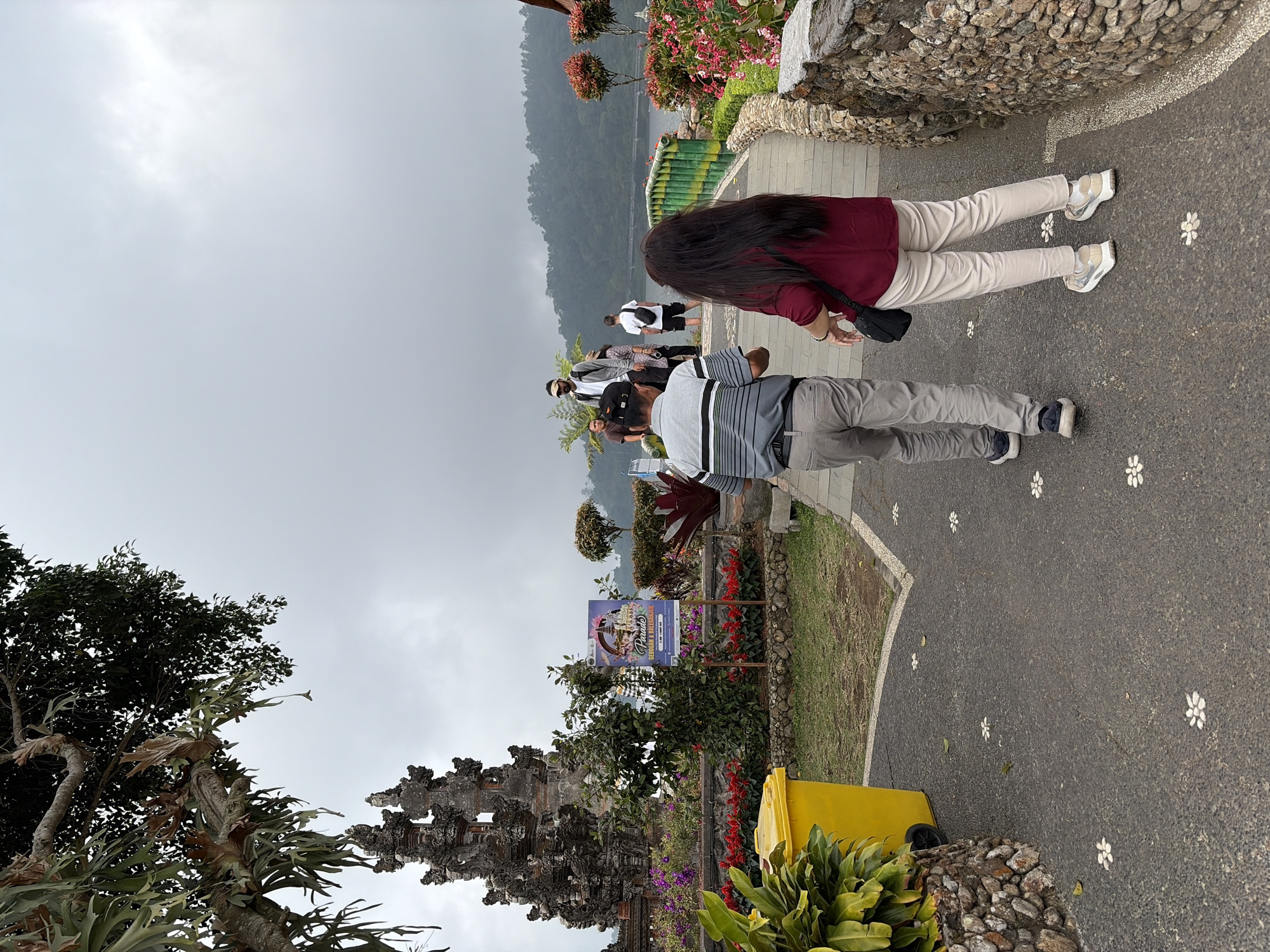 This image captures a scene at Pura Ulun Danu Bratan, a Hindu temple complex located in Baturiti, Bali, Indonesia. In the foreground, a couple walks away from the camera on a paved pathway decorated with white flower-like inlays. The man, on the left, wears a gray and black striped polo shirt and light gray pants. The woman, on the right, is dressed in a maroon shirt and light beige pants. Behind them, other visitors are visible on a bridge that spans over water, with lush green hills and a lake in the background. To the left, a large, ornate stone temple structure stands prominently, adorned with intricate carvings. Bright yellow flowers and greenery surround the base of the temple, and a bright yellow trash can is situated near the pathway. The sky is overcast, contributing to a diffused, even lighting across the scene.