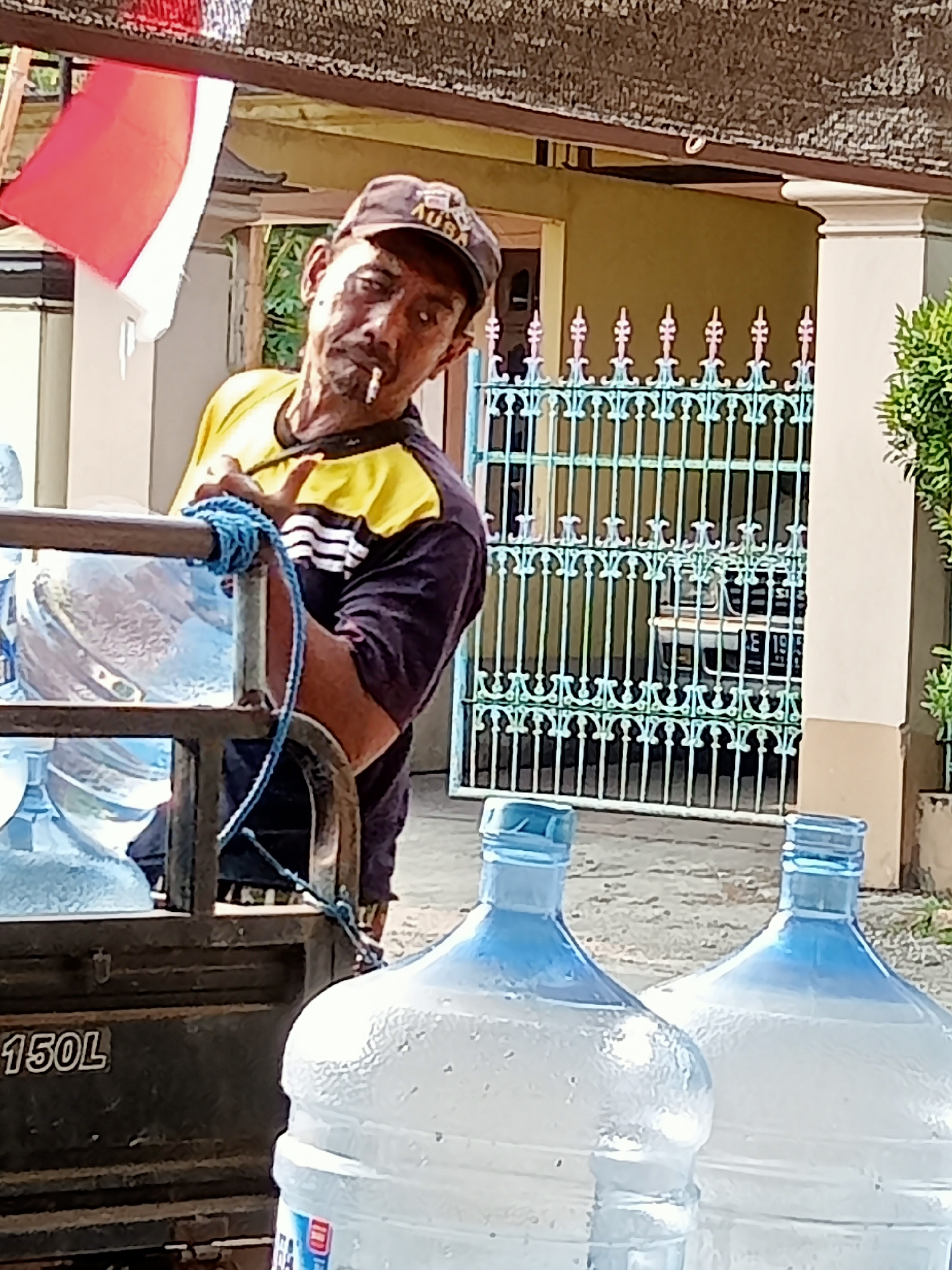 The image depicts a man smoking a cigarette, with a blue rope tied around his wrist and the frame of a vehicle. He is wearing a yellow and dark blue shirt and a dark cap. The vehicle appears to be a truck with "150L" visible on its side, carrying multiple large water jugs. In the background, there is a house with a white pillar and a decorative metal fence. An Indonesian flag is visible on the left side of the frame. The scene is outdoors, possibly in the city of Simo, Indonesia, suggested by the presence of the Indonesian flag.