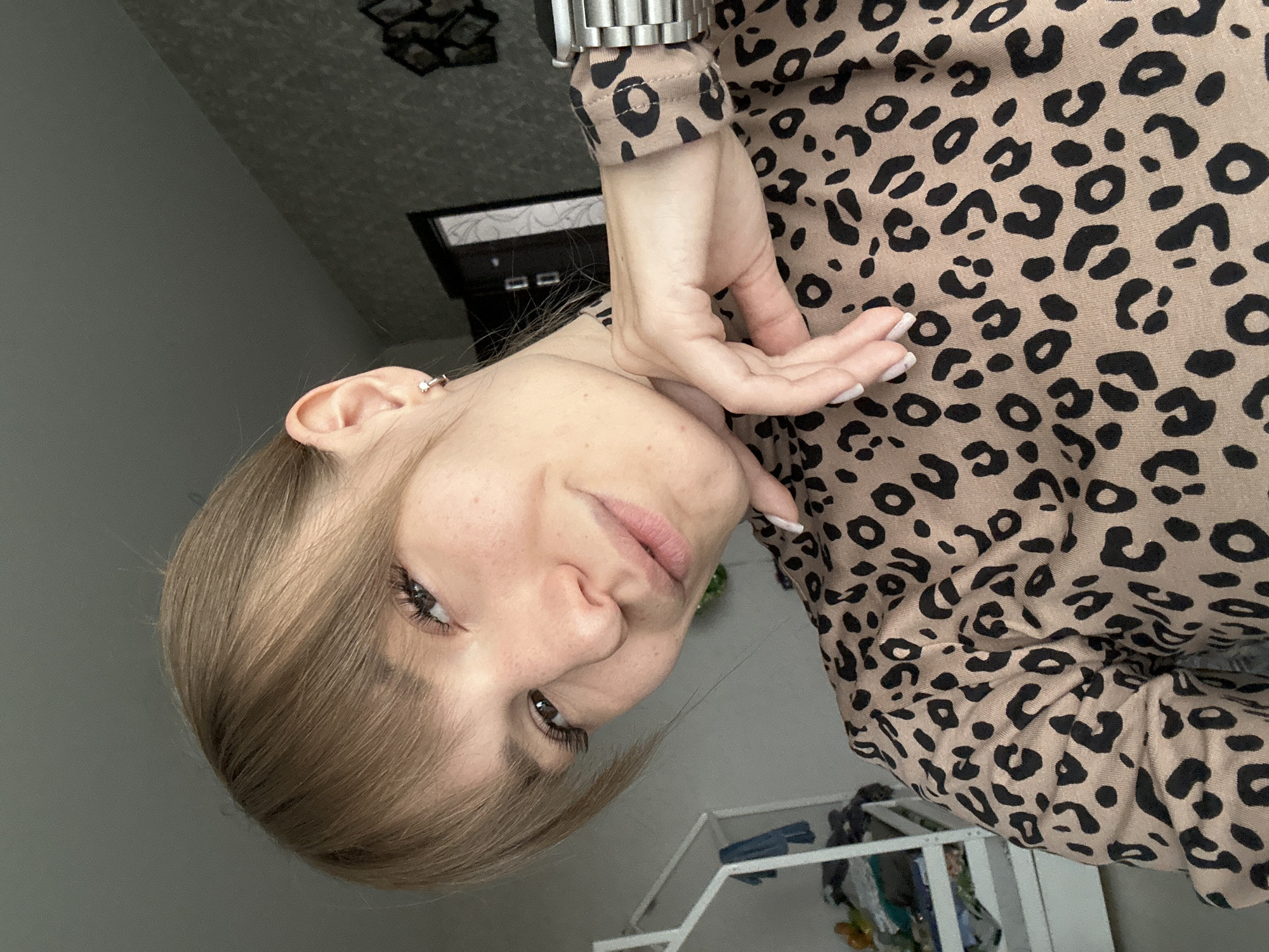 A young woman with blonde hair and bangs is taking a selfie indoors. She is wearing a leopard-print top and a silver watch. Her left hand is resting on her chin, and she is looking at the camera with a slight smile. The background appears to be a bedroom with a white crib and some clothing on it. The room has grey walls and a door in the background. The image was taken on March 10, 2021, in Chaikovsky, Russia.