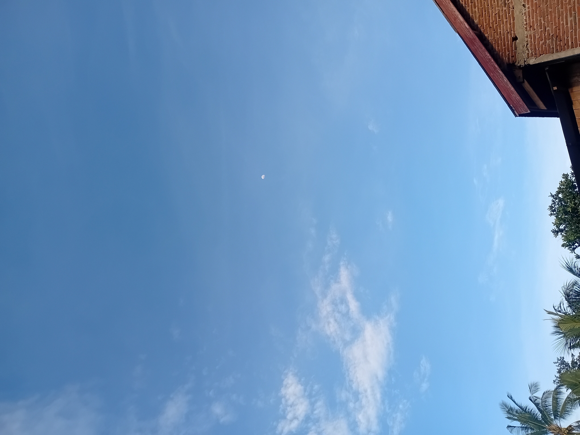 A clear blue sky with scattered white clouds dominates the upper portion of the image. A small, crescent moon is visible in the upper center. The bottom of the image features the roof and brick wall of a building on the right, with the tops of palm trees and other foliage on the left. The scene appears to be an outdoor view from Lhokseumawe, Indonesia.