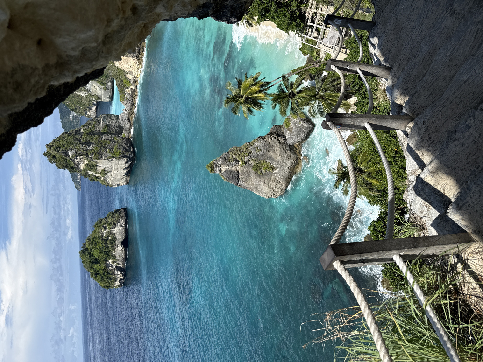 The image depicts the landscape of Nusa Penida, a small island southeast of Bali, Indonesia, specifically in the city of Pejukutan. The view is from a high vantage point, looking down over the coast. Turquoise water dominates the scene, contrasting with rocky islands covered in greenery. A white sand beach is visible in the distance. The image is taken from what seems like a cave or natural overhang, with a rocky wall on one side, and a man-made rope fence is present on the other. The bright sunlight casts long shadows.