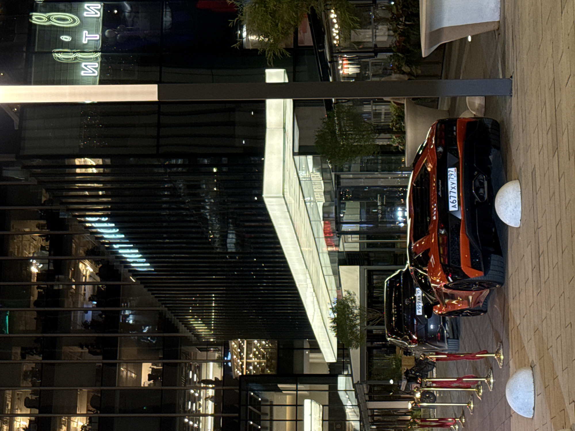 A nighttime, low-angle shot of two sports cars parked along a brick-paved area in Khimki, Russia. The first vehicle, a black and orange Lamborghini, has a license plate number A677XY797. The second, a black SUV, features the license plate P999AA95. A row of gold stanchions with red ropes lines the side of the cars, guiding pedestrians along the brick surface. The architecture consists of modern, glass-fronted buildings. Light reflects off the glass surfaces. Plants are situated on the facade of the buildings. Street lighting and reflections create a complex interplay of light and shadow. The scene is well-lit and appears to be a high-end or luxury location.