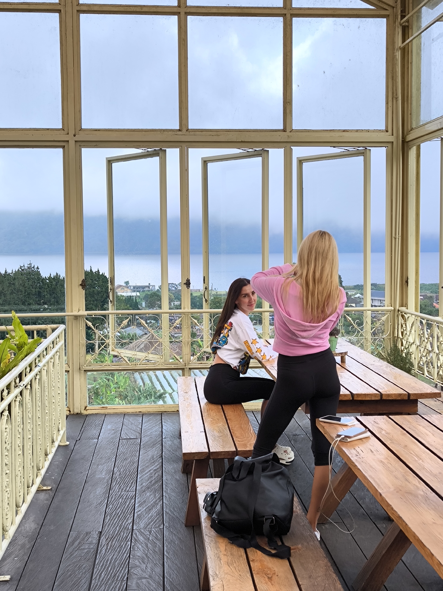 Inside a sunroom in Candikuning, Indonesia, two young women are present. One, with long dark hair, is seated on a wooden bench facing the camera, while the other, with long blonde hair, stands next to a wooden table with her back towards the viewer. The blonde is adjusting the hood of her pink sweatshirt. The seated woman is wearing a white long-sleeved shirt with colorful graphics, paired with black leggings.
The sunroom has a wooden floor and a series of large windows, some of which are open, providing a view of a body of water, a town, and a mist-covered sky. Additional details include a black backpack on a bench, and two mobile phones and a charging cable on the table next to the blonde woman. Metal railings are visible outside the windows.