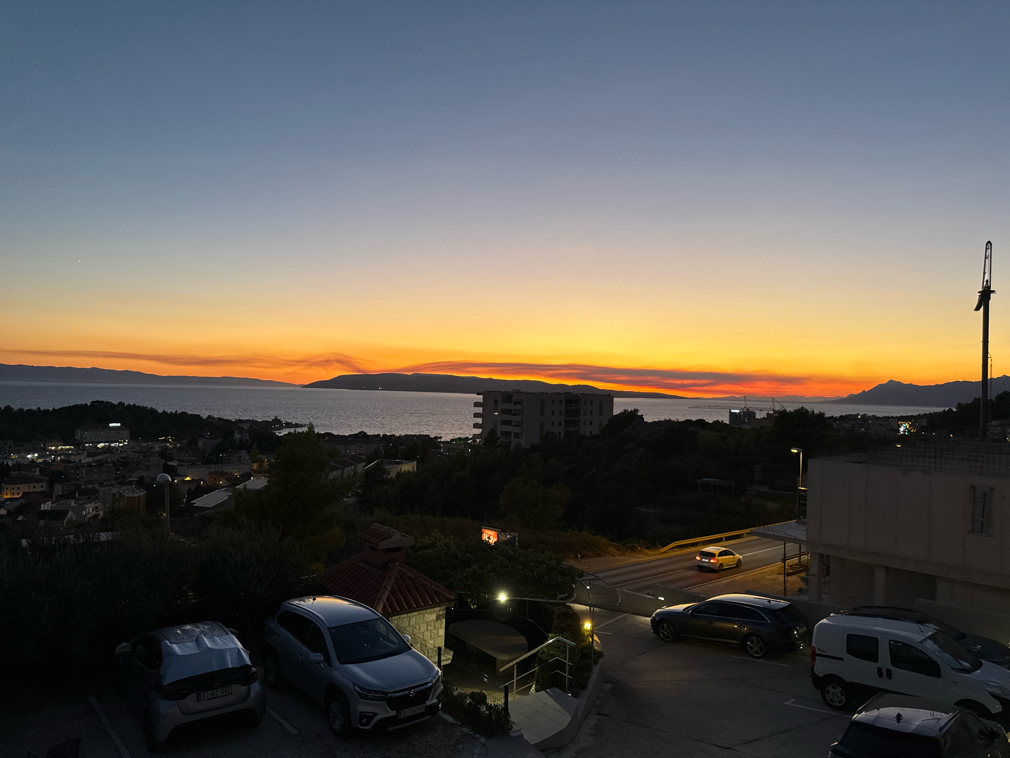 The image captures a sunset over the Adriatic Sea, viewed from a slightly elevated position within the city of Makarska, Croatia. In the foreground, several parked cars are visible, including a silver hatchback with license plate "AI-KC-080" and a silver SUV with license plate "ZG 3524 KA". Behind the cars, there are stairs leading down to a courtyard area. Further into the midground, the town of Makarska unfolds with numerous buildings and streetlights illuminated against the darkening sky. A large, modern building with multiple stories stands prominently overlooking the sea. The sea itself is calm, with a distant mountain range silhouetted against the vibrant orange and yellow hues of the sunset. The sky transitions from a deep blue at the top to a gradient of orange and yellow near the horizon. A tall construction crane is visible in the distance on the left side of the image. A few vehicles are driving on a road in the midground, their headlights illuminating the scene. The overall atmosphere is one of twilight transitioning into night, with the natural beauty of the sunset juxtaposed with the urban landscape.