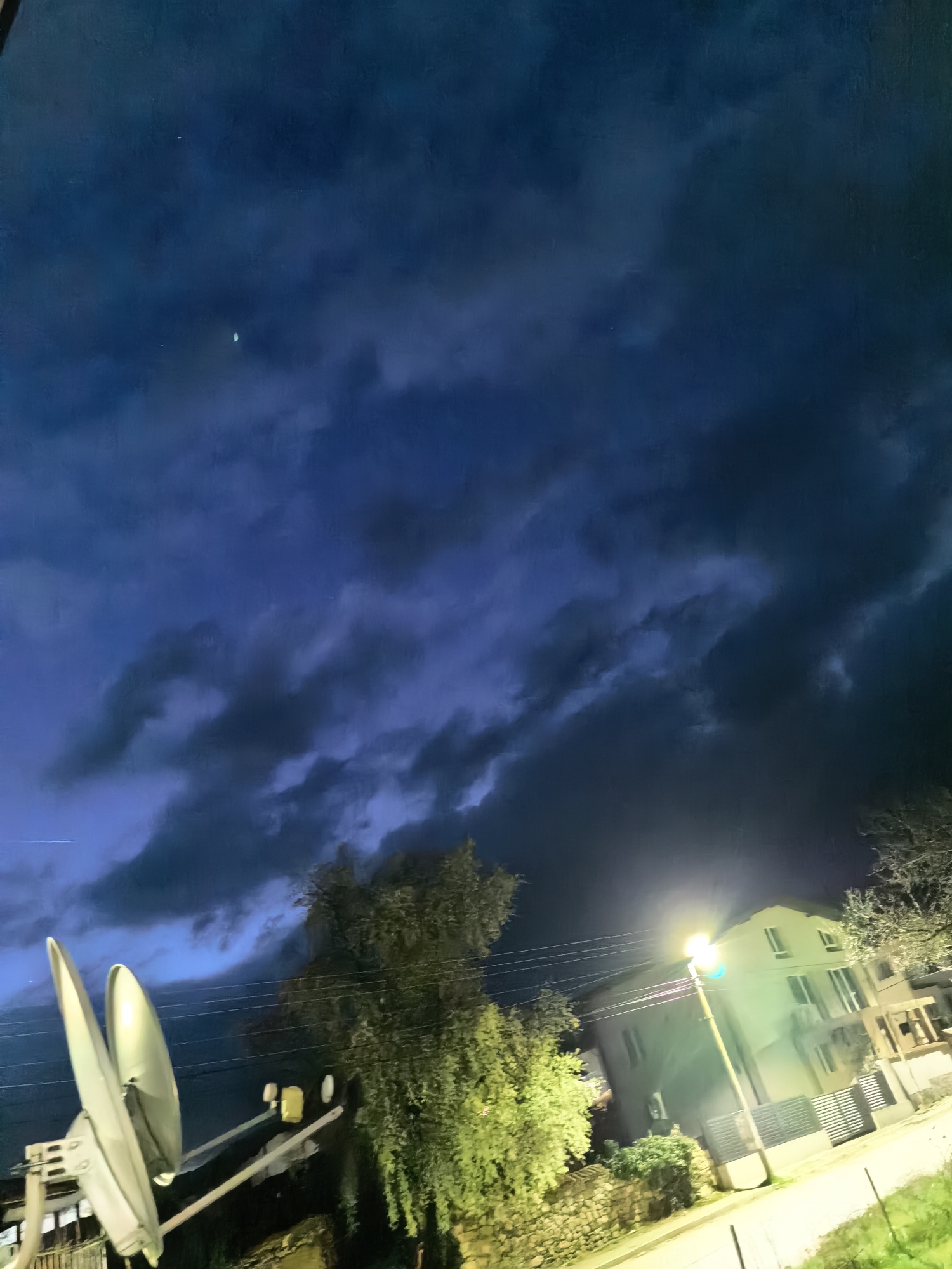The image presents a nighttime scene in an urban location. The sky is dominated by dark, turbulent clouds, with what appears to be a faint star visible. In the lower portion of the image, there is a satellite dish on the left. A tree with green foliage stands near the center of the image. In the background, there is a building illuminated by a street lamp, which casts a bright glow. The scene is captured from ground level. The location is in an urban area.