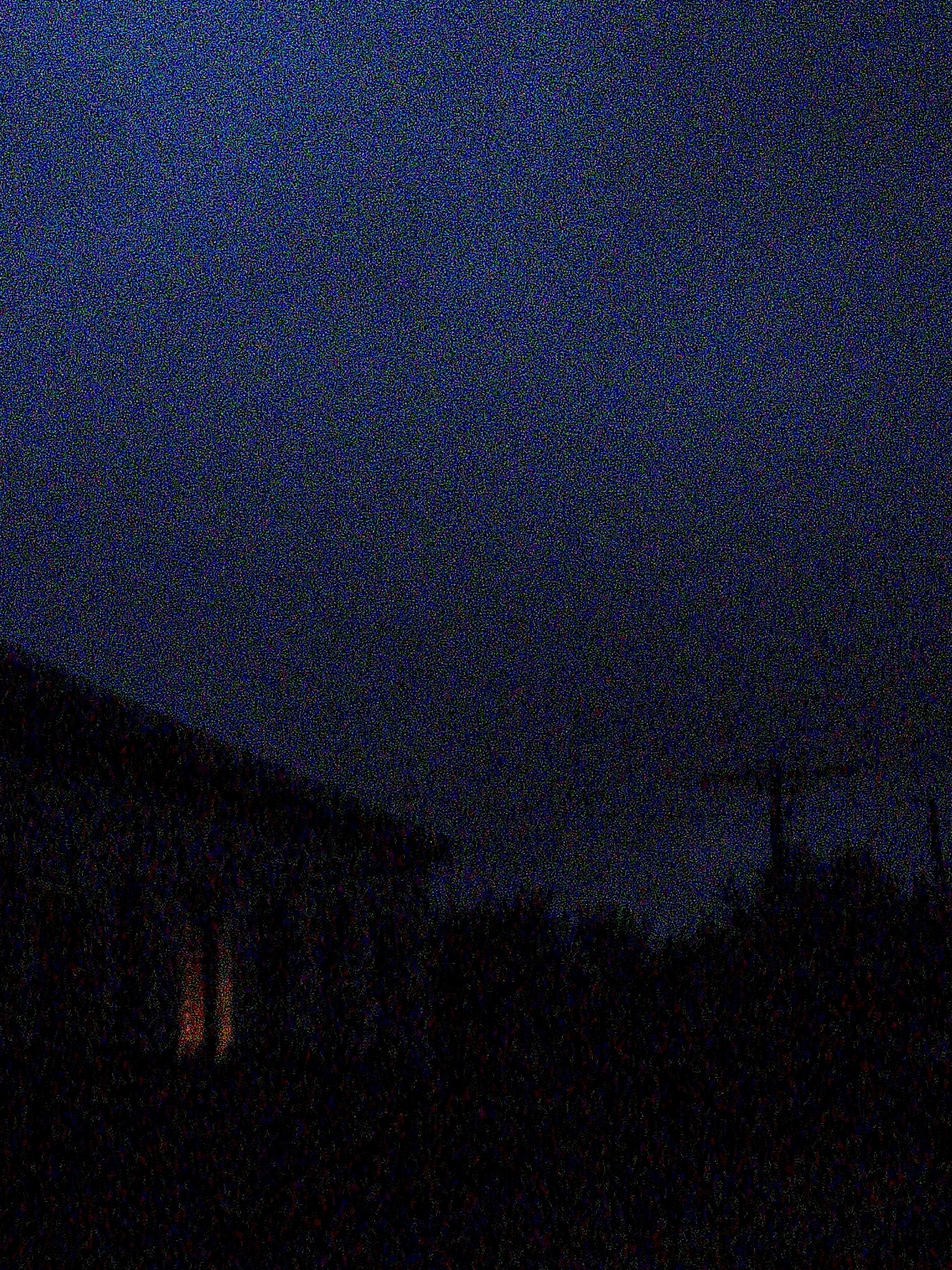 The image depicts a nighttime scene with a dark, grainy texture. In the foreground, a dark, silhouetted structure, possibly a building or roofline, is visible on the left side. Two vertical, reddish-orange lights are emanating from within this structure, suggesting interior lighting. The rest of the foreground is obscured by darkness and appears to be foliage or trees. In the background, the sky is a deep, dark blue, with a faint hint of a structure, possibly a utility pole or tower, silhouetted against the sky on the right. The overall impression is of a quiet, possibly rural or suburban, nighttime setting. The provided context of Ustyvytsia, Ukraine, indicates the geographical location of this scene.