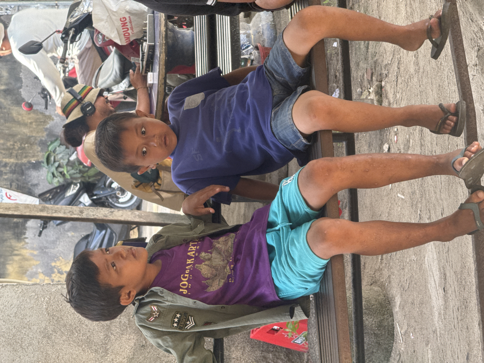 Two young boys lie sprawled on a metal bench in Nusa Penida, Indonesia. The boy on top wears a blue t-shirt and denim shorts, while the boy below sports a purple t-shirt, turquoise shorts, and a green army-style jacket. Both wear flip-flops. In the background, a man with a Rasta hat and headphones stands near a motorbike. A plastic bag labeled "ANDUNG Collection" hangs from the motorbike. Debris litters the ground around the bench and the gray concrete wall behind the bench.