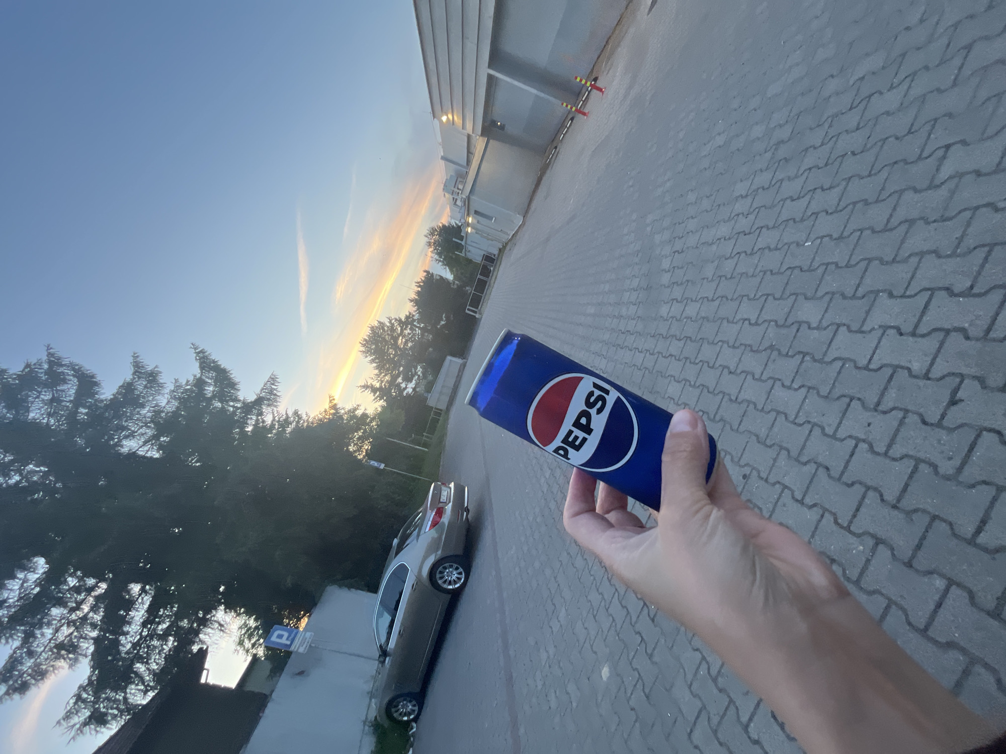 A hand holds a can of Pepsi, its logo prominently displayed, in an outdoor setting with a paved walkway. In the background, a light-colored sedan is parked. The environment features a building with a modern facade and some trees. The sky is a clear blue with streaks of orange and pink clouds, suggesting either sunrise or sunset. A parking sign is visible on a post near the building.