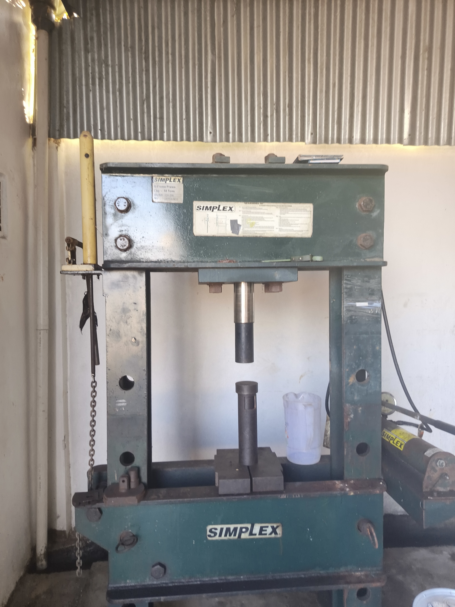 The image shows a green hydraulic press machine, labeled "SIMPLEX," positioned in what appears to be an outdoor workshop or garage setting. The machine is centrally located and occupies a significant portion of the frame. To the left of the machine, a corrugated metal wall or roof is visible, with a yellow vertical pole standing in front of it. A chain hangs from the left side of the press. In the background, a light-colored wall is present. On the right side of the press, a hydraulic cylinder is attached, also labeled "SIMPLEX." A clear plastic container, possibly a pitcher or jug, is placed on the work surface of the press. The ground beneath the machine appears to be concrete. There are no people or animals visible in the image. The scene suggests a functional industrial or mechanical workspace.