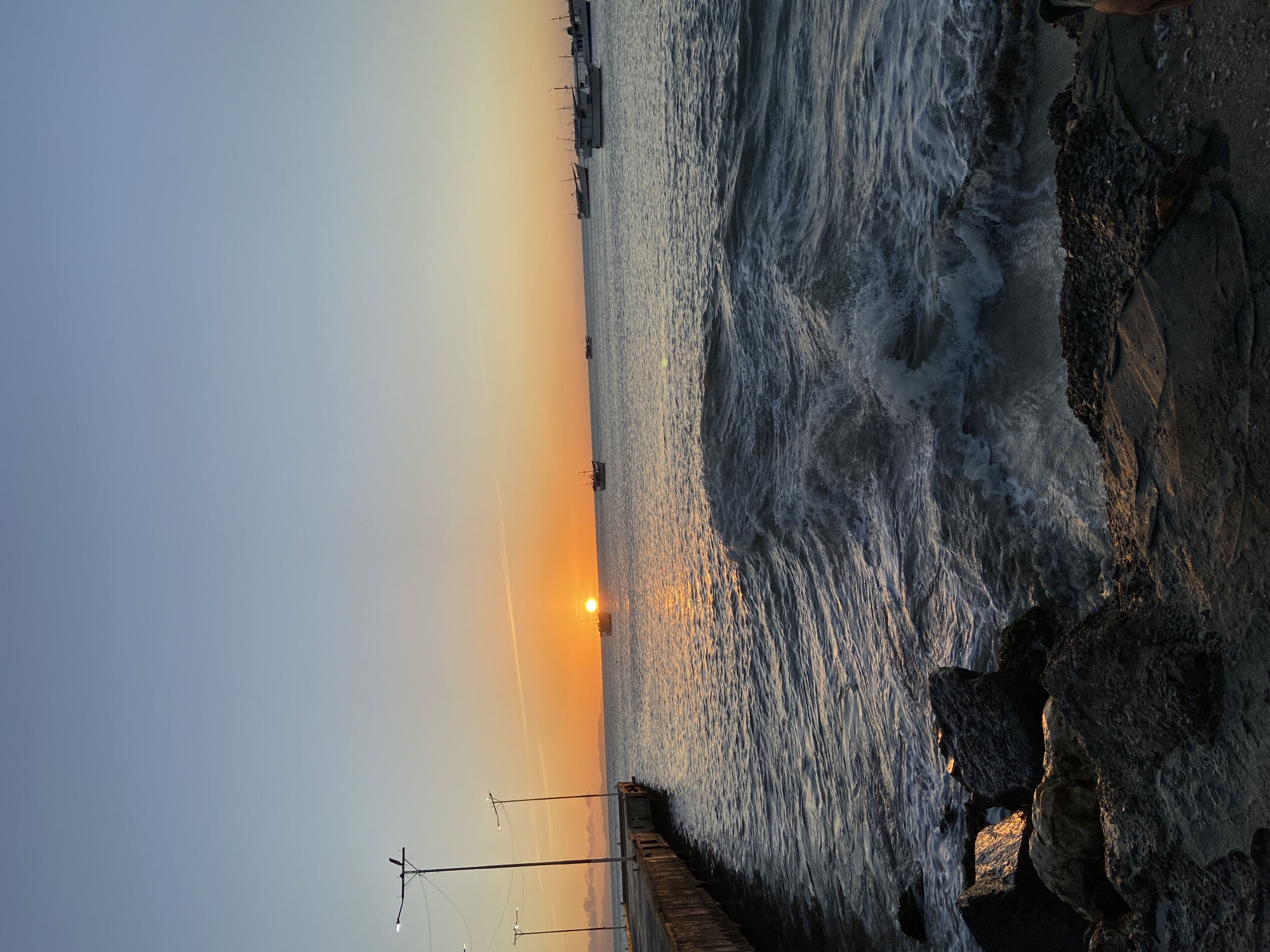 The image shows a sunset over the ocean in Juan Griego, Venezuela. The sky transitions from blue to orange as the sun nears the horizon. Several boats are silhouetted in the distance. The ocean is choppy with visible waves reflecting the sunlight. A rocky shoreline is on the right side of the image. Several telephone poles extend from a structure on the left side of the image.