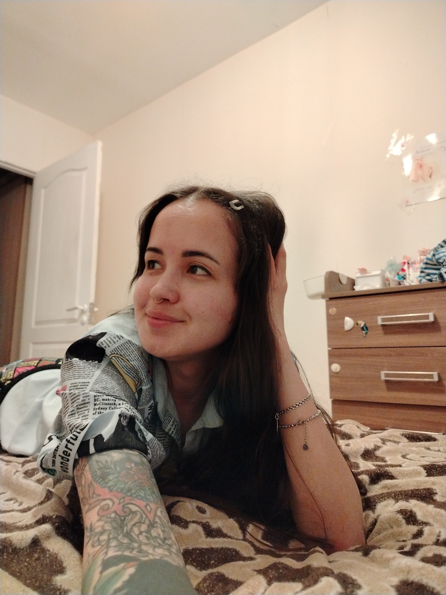 A woman with long dark hair is lying on a bed covered with a brown and tan patterned blanket. She is resting her head on her left hand, which has a bracelet on the wrist. Her left arm, which is heavily tattooed, is extended towards the viewer. She is wearing a light-colored shirt with a newspaper print design on the sleeves. She is looking off to the left with a slight smile.
In the background, a white door is partially visible on the left, and a brown dresser with two drawers is on the right. On top of the dresser are various containers and objects. The walls are painted a light beige color.