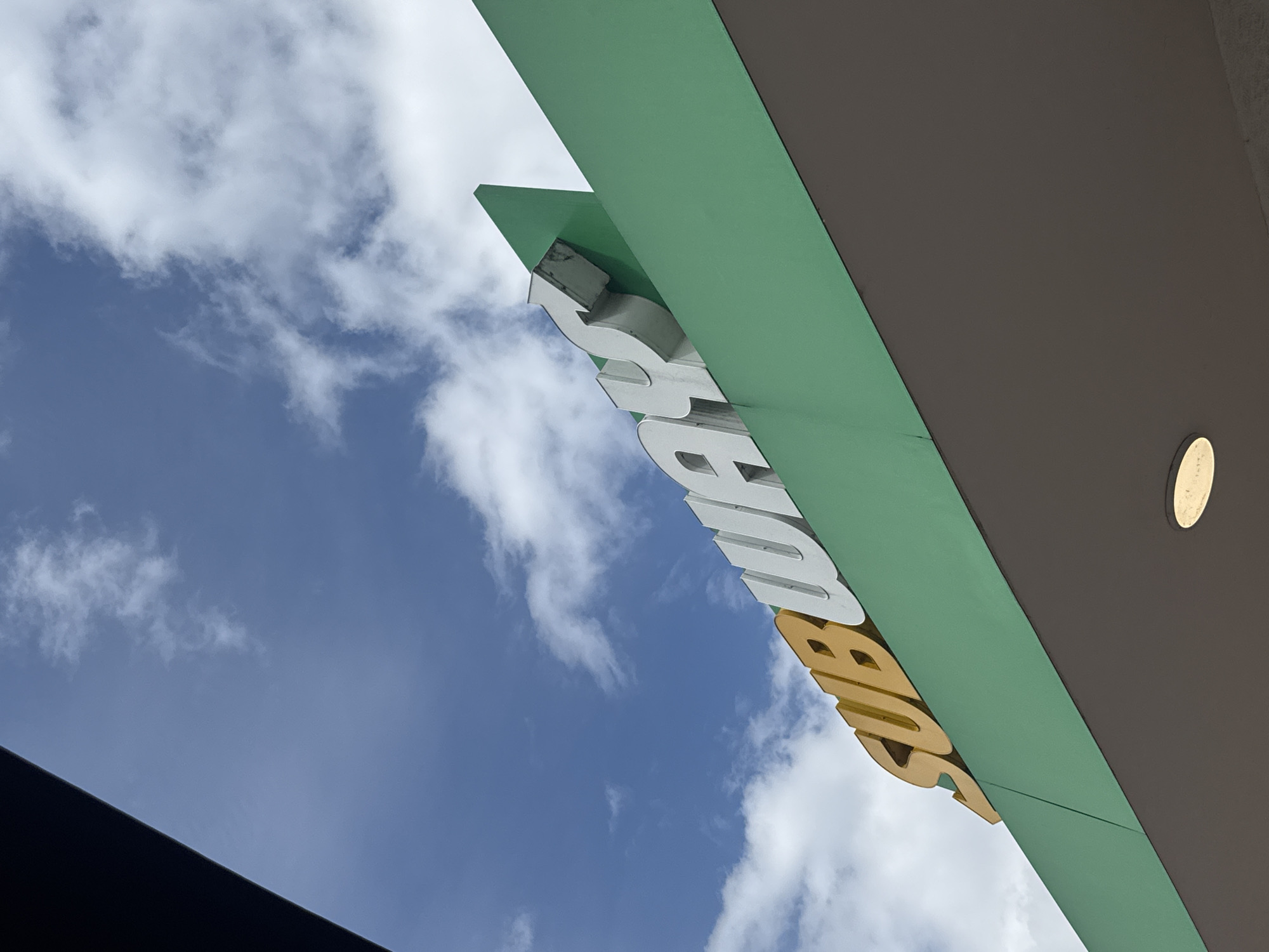 An angled outdoor shot shows a green building overhang with a "SUBWAY" sign. The sign is white and yellow with white letters that spell out the brand name. The sky behind the sign is blue with scattered white clouds. The lower portion of the image is a dark grey overhang with a light fixture.