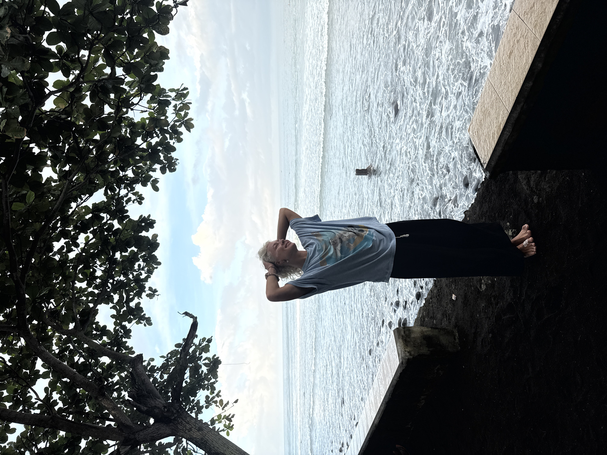 A medium shot shows a Caucasian woman standing on a beach in Unknown, Unknown. The woman has short, white hair and is wearing a blue t-shirt and black pants. She is standing on the edge of a concrete structure that borders the beach. The ocean is behind her, with waves crashing on the shore. A tree with green leaves is to the left of the woman. The sky is cloudy and overcast. The woman is looking up and has her hands behind her head.