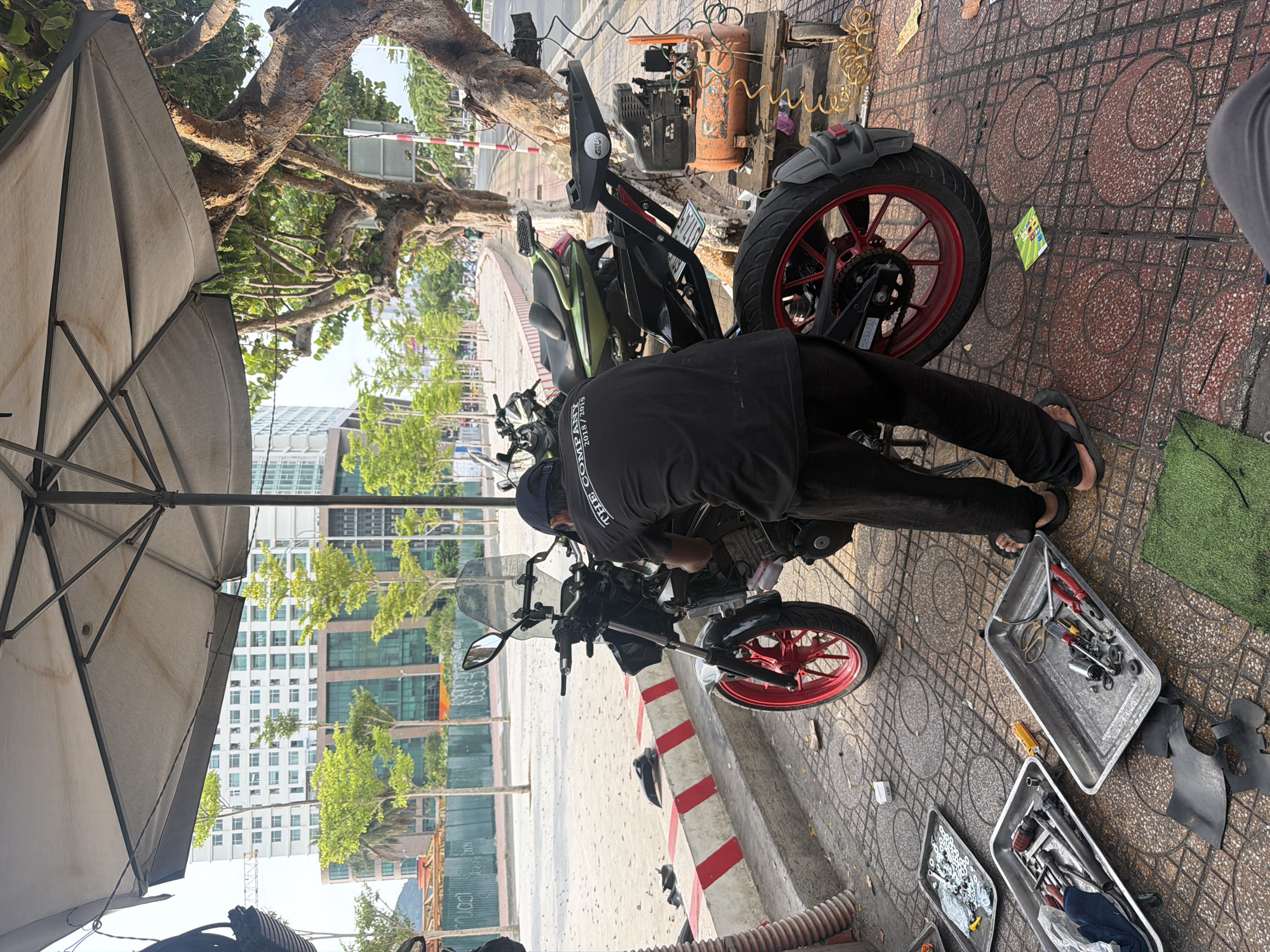 In Nha Trang Ward, Vietnam, a mechanic is working on a motorcycle under a large umbrella. The mechanic, wearing a black t-shirt and black pants, is positioned behind the motorcycle, appearing to adjust or repair it. Various tools are laid out on the ground in metal trays, suggesting a repair session is underway. A small orange air compressor on a wooden cart is situated to the right of the motorcycle, indicating potential use for tire inflation or other pneumatic tasks. In the background, modern buildings and trees line a street, characteristic of an urban environment. The scene is set on a paved sidewalk with red and white striped curb detailing.