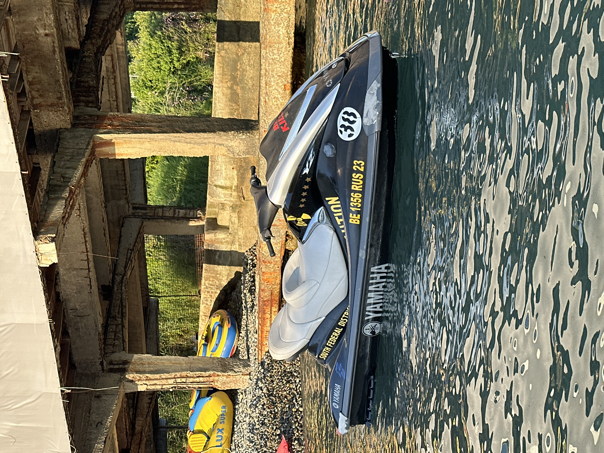 A black Yamaha jet ski is floating on calm, clear water. The jet ski has a white seat and is branded with "YAMAHA" in large white lettering. It also has a sticker that reads "SOUTH FEDERAL DISTRICT" and a license plate that says "BE 1356 RUS 23." The jet ski is positioned in front of a concrete structure with visible rebar, suggesting it may be an abandoned or under-construction pier or building. In the background, there are two inflatable water tubes, one yellow and blue labeled "LUX SUPER," and lush green vegetation. The scene appears to be located in Sochi, Russia, given the license plate information. The lighting suggests it is either early morning or late afternoon, with warm sunlight casting long shadows. No people are visible in the image.