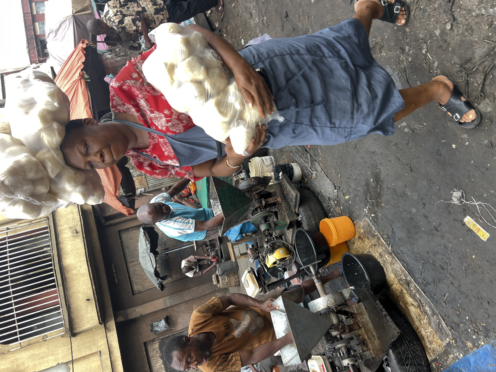 The image depicts a bustling street scene in Nkpor, Nigeria, during daylight hours, likely within a market or an informal industrial area.
In the foreground, a woman is centrally positioned, balancing a large bag of white, spherical items on her head while also carrying another similar bag in her arms. She wears a red floral-patterned top over a blue apron or dress and black sandals. Her gaze is directed towards the viewer, with a calm expression.
Behind her, several men are engaged in operating machinery. One man, wearing a light blue and white striped polo shirt, stands beside a green processing machine. Another man, closer to the foreground, is dressed in a brown t-shirt patterned with "DIOR" and is actively feeding material into a metal-hoppered machine, suggesting a milling or grinding operation. Additional figures are visible in the background amidst various stalls and structures.
The ground is uneven and appears to be a mix of dirt and worn concrete, scattered with debris including tire fragments, wires, and plastic. Orange and black plastic buckets are placed near the machinery. The surrounding structures include buildings with barred windows and simple awnings, indicative of a busy commercial or workshop setting. The scene conveys a sense of industrious activity in a vibrant, functional environment.