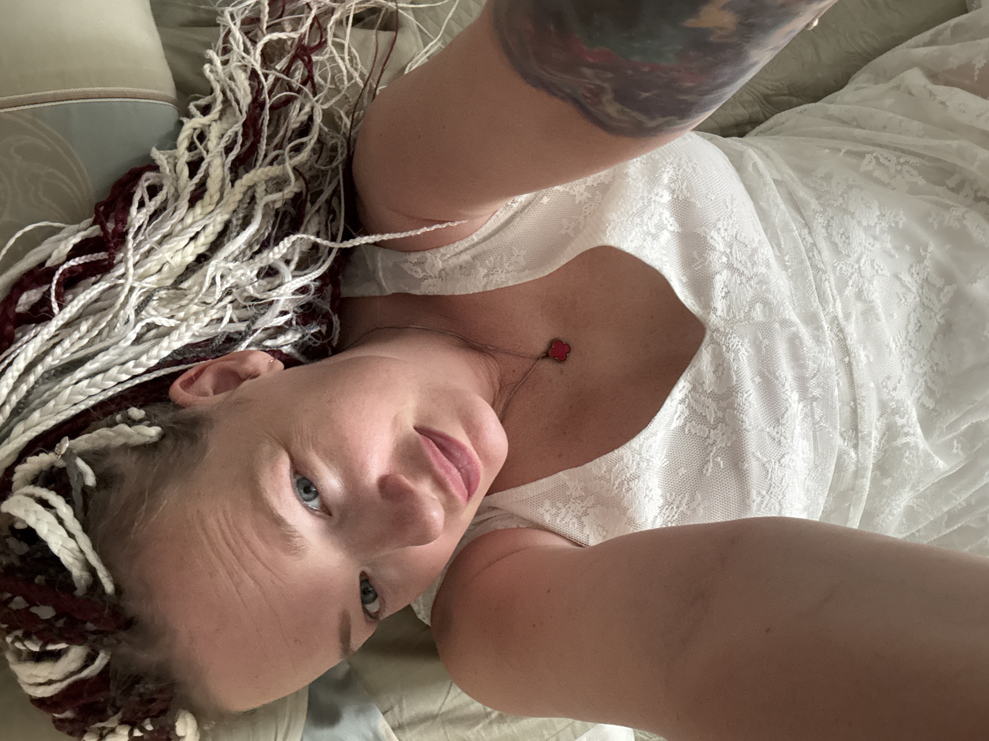 A woman with braided hair in shades of white, cream, and burgundy lies on her side on a bed. She is wearing a white lace dress. Her left arm is extended and angled upwards, with a tattoo visible on her upper arm. A delicate necklace with a small red heart pendant rests on her chest. The woman's eyes are open and she is looking towards the camera with a slight smile. Her facial expression suggests a relaxed and intimate mood. The background consists of pillows and bedding, hinting at a domestic setting. The lighting appears soft, possibly indicating natural light from a window, suggesting it could be daytime. There is no visible text in the image. Given the location context of Moscow, Russia, the scene is likely indoors in a bedroom.