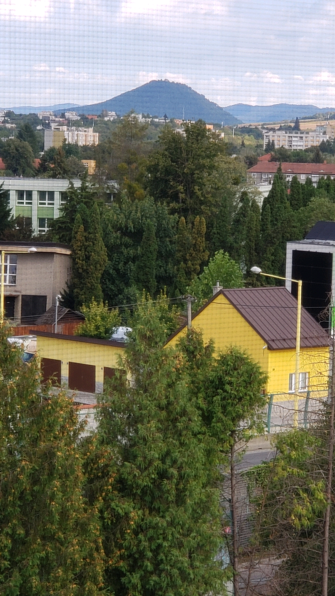 This is a photograph taken from an elevated perspective, likely from a window, looking out over a cityscape with a prominent mountain in the background. The foreground features lush green trees, some with yellowing foliage, obscuring parts of the buildings below. A yellow house with a brown roof and a prominent streetlamp in front of it is visible. Adjacent to it is a lighter-colored building with garage doors. Further back, more trees are interspersed with various urban structures, including residential buildings and what appears to be a school or institutional building with green accents. In the distance, a distinctive cone-shaped mountain, likely the Zemplin Mountains, dominates the skyline, with other hilly terrain and scattered buildings extending towards it. The sky is partly cloudy. The scene depicts a residential or mixed-use area within the city of Prešov, Slovakia, with a natural landscape in the background.