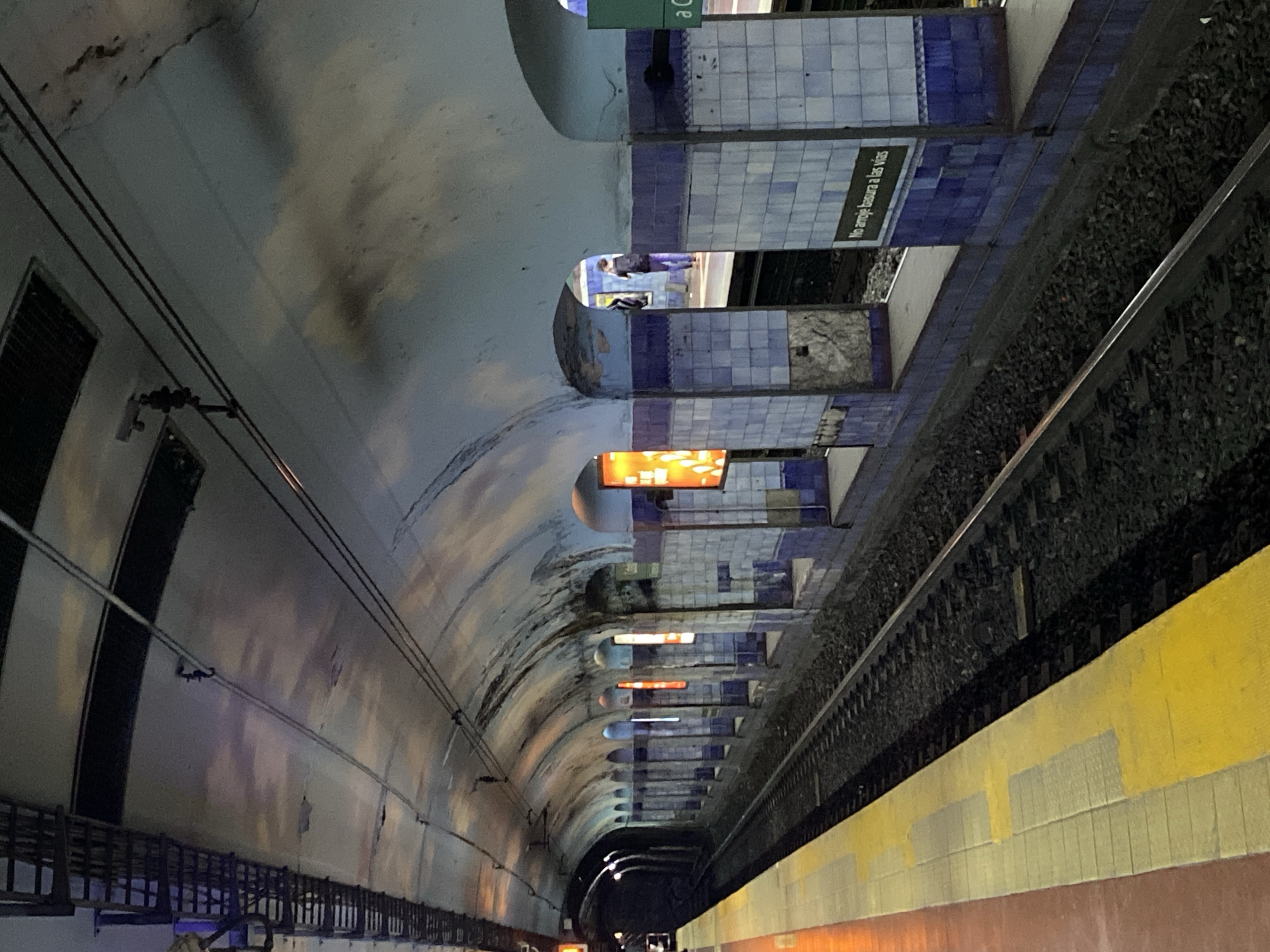 Here's a description of the image:
This image depicts an underground metro station in Buenos Aires, Argentina. The station platform is lined with yellow and red painted tiles. The adjacent subway tracks are filled with ballast. Above the tracks, sections of wall are decorated with blue and white tiling. The tunnel ceiling curves overhead, revealing a series of arched openings along the platform edge, each housing illuminated signs. In one archway a person is visible on the opposite platform. The overall impression is that the station is aged and in need of repair.