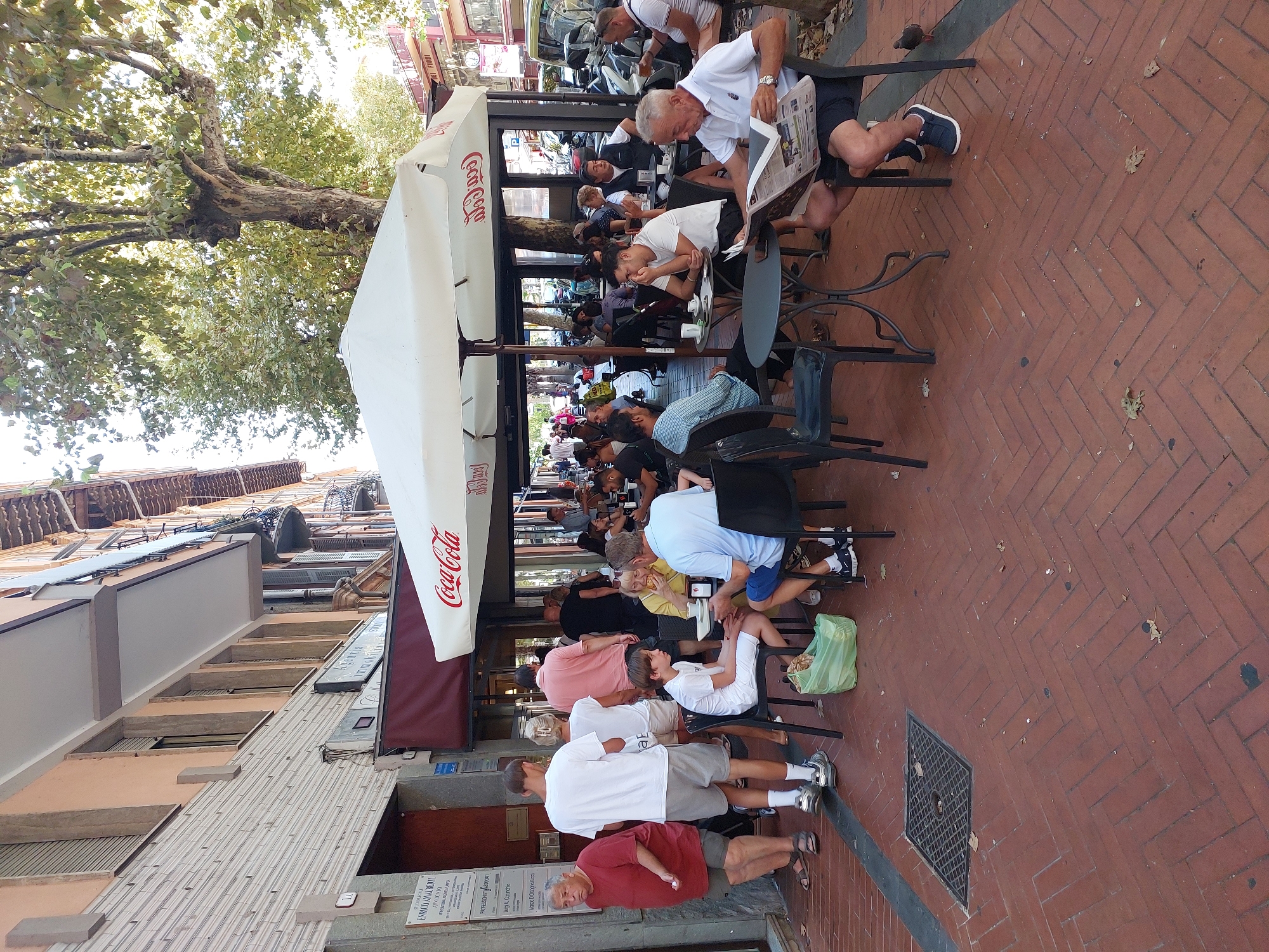 This image depicts an outdoor cafe scene on a sunny day, likely in Ventimiglia, Italy, given the architectural style and ambiance. The foreground is dominated by a red brick herringbone-patterned sidewalk. A large white umbrella with "Coca-Cola" branding shades a seating area with several tables and chairs.
Several individuals are present, either seated at tables or standing. In the foreground, an elderly man wearing a light blue polo shirt and dark shorts sits at a table, engrossed in reading a newspaper. To his right, a person in a dark blue shirt and shorts is seated, with a portion of their leg visible. Further back, a young boy in a white t-shirt and shorts sits at a small table. To his left, a woman in a yellow top is seated, appearing to eat.
Behind the main seating area, more people are visible at tables, some conversing and others engaged with their food and drinks. A group of people are standing near the entrance of a building to the left. One man in a red t-shirt and shorts is standing prominently, looking to his left. Two younger men in white t-shirts and shorts are also standing.
The background shows the facade of a building with multiple windows and balconies, some with shutters. Trees with lush green foliage overhang the scene, casting dappled shadows. In the distance, the street is visible with parked vehicles and other pedestrians. The overall impression is one of a bustling, relaxed atmosphere typical of an Italian piazza cafe.