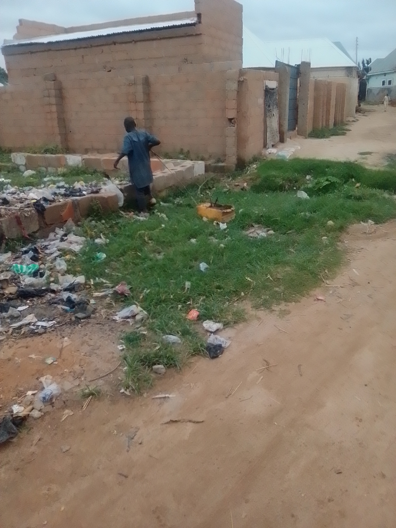 A young Black male in a dark blue t-shirt and dark pants stands on a dirt path in front of a brick building with a corrugated metal roof. He is facing away from the camera, with his left arm extended and holding a stick, as if sweeping or clearing debris. In the foreground, to the left of the male, is a pile of trash, including plastic bags, bottles, and other refuse, accumulated around a low-lying brick structure. The ground is mostly dirt and scattered with some green grass and more trash. In the background, to the right of the building, are other structures with similar roofing materials, suggesting a residential or commercial area. The sky is overcast. The location is the city of Gombe, Nigeria.