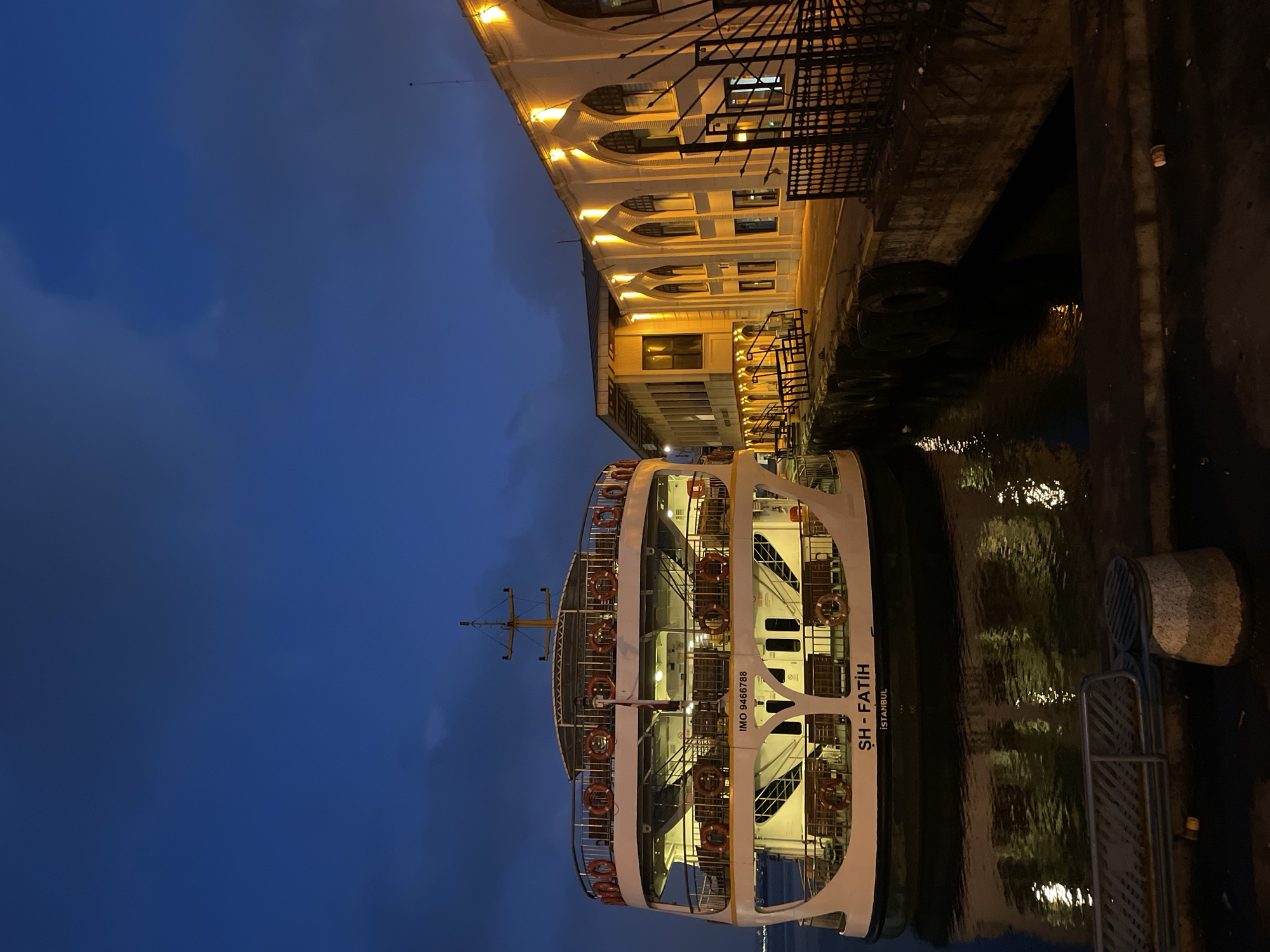 The image depicts a ferry docked at a pier in Kadıköy, Turkey, during twilight. The ferry, named "ŞH - FATİH," is white with multiple decks and adorned with orange life preservers. It is illuminated from within, casting a warm glow onto the water. The pier features a long, white building with arched windows, also lit from within, suggesting it is a terminal or administrative building. The water reflects the lights of the ferry and the building. The sky is a deep blue, with some clouds visible. The scene appears calm and serene, with no people visible. The overall impression is one of urban maritime activity at the close of day.