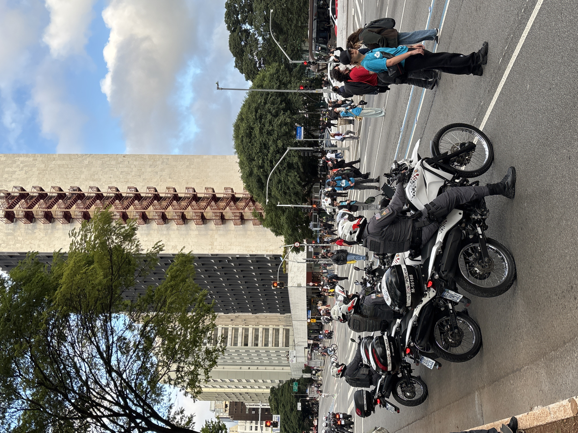 The image depicts a street scene in São Paulo, Brazil. A row of parked police motorcycles dominates the foreground, facing toward the center of the frame. Several officers are seen sitting atop the motorcycles, facing the same direction. The bikes have black and white color schemes with motorcycle helmets strapped in place.
Behind the motorcycles, a crowd of people waits at a crosswalk. Some of them are mid-stride, walking across the street. Several red traffic lights are visible overhead.
In the background stands a tall building with an external fire escape. Trees line the street, obscuring some of the buildings. The sky is partly cloudy with patches of blue.