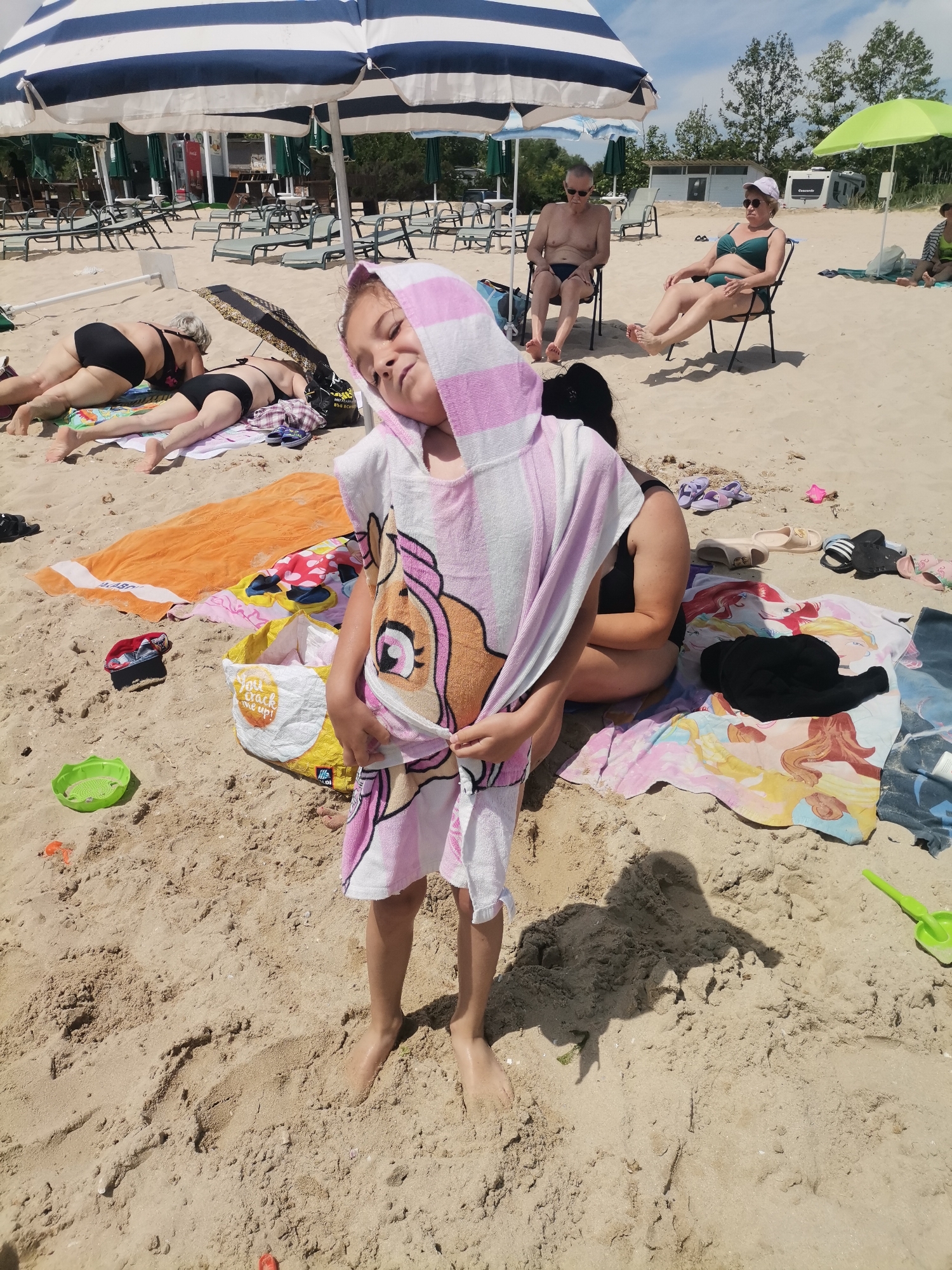 The image captures a sunny day at Kranevo beach, Bulgaria, with a focus on a young girl in the foreground. She is wearing a pink and white striped hooded towel with a cartoon character design. Her head is tilted, and she appears to be looking towards the camera with a playful expression. Around her, the sandy beach is populated with other beachgoers. To the left, two women are lying on towels under a large blue and white striped umbrella. Further back, a man and a woman are seated in chairs under umbrellas. Various beach accessories are scattered across the sand, including towels, bags, flip-flops, and a green plastic shovel. The background shows more beach chairs, umbrellas, and some vegetation. The overall scene depicts a relaxed atmosphere on a crowded beach.