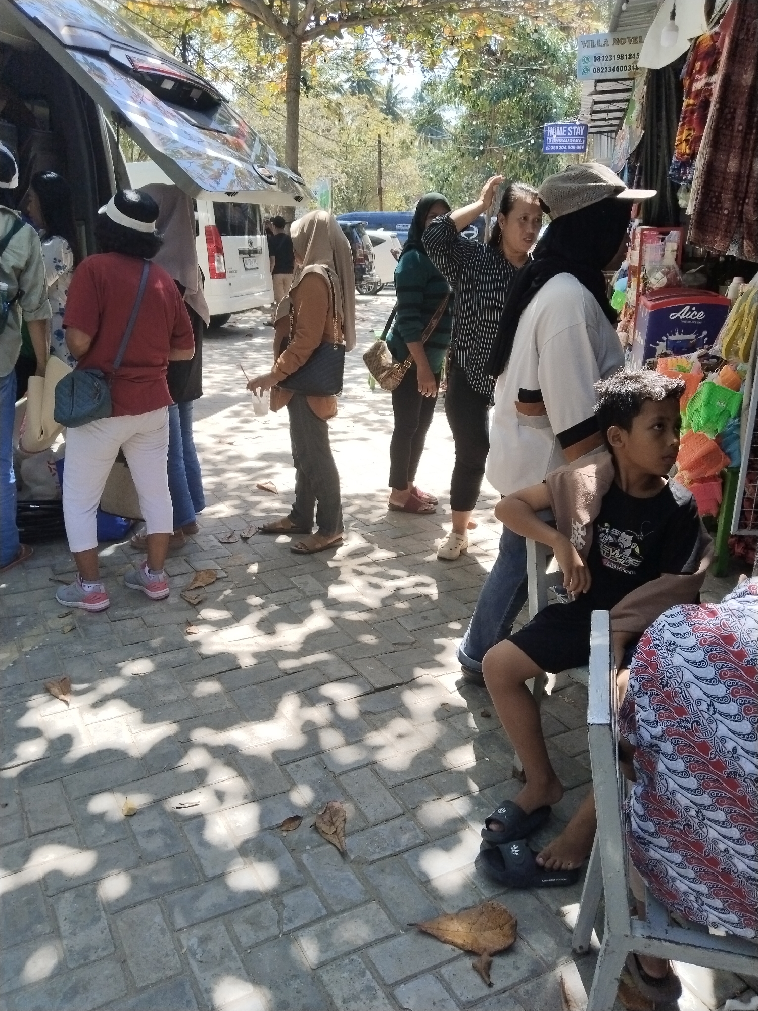 The image depicts a street scene in Prigi, Indonesia, likely near a local market or roadside establishment. In the foreground, a young boy with dark, spiky hair sits on a white metal chair, wearing a black t-shirt and black shorts. He is looking to his right. To his left, a person wearing a red shirt and white cap is visible from the chest down, with a blue bag slung over their shoulder. Several other individuals are present, some standing and others partially obscured, including a person in a brown hijab and another in a black hijab.
Behind the people, a white van with its rear doors open is parked. The van appears to be a common model for passenger transport. To the right of the van, a stall displays clothing and other merchandise, with a blue and white cooler labeled "Aice" visible. A sign above the stall reads "VILLA NOVEL" and "HOME STAY." The ground is paved with grey interlocking bricks, and scattered dry leaves are present, suggesting an outdoor environment with some natural elements. Sunlight filters through unseen trees, casting dappled shadows on the pavement, indicating a sunny day. The overall impression is of a bustling, informal commercial area in Indonesia.