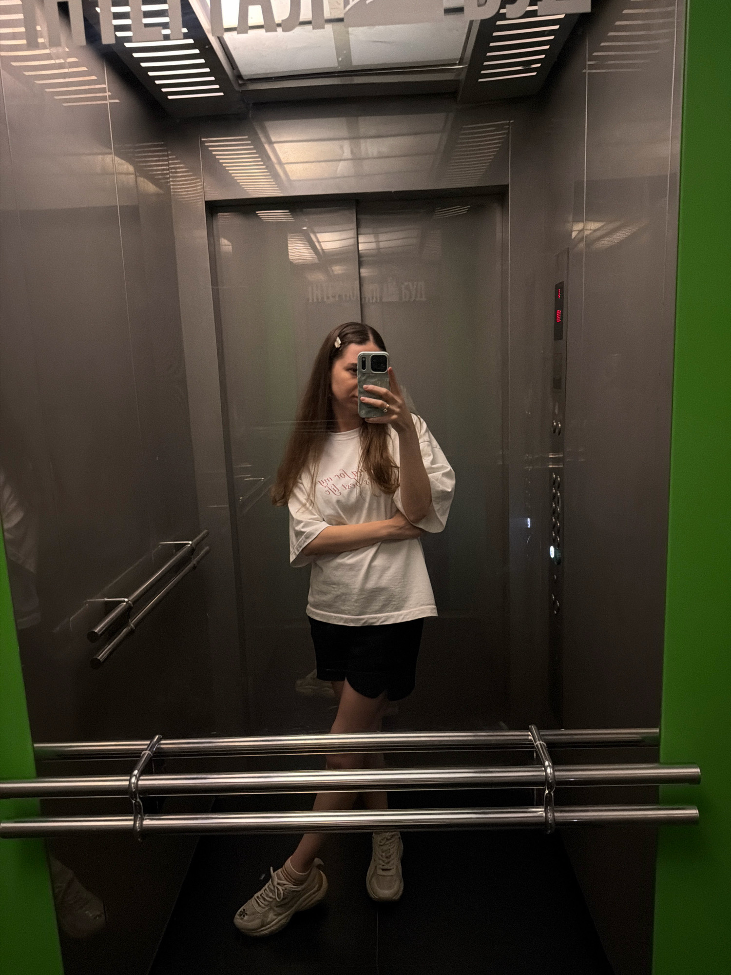 The image shows a young woman taking a mirror selfie inside an elevator. She is wearing a white t-shirt with pink lettering and black shorts, paired with white sneakers. She has long brown hair and is holding a smartphone in her right hand, with her arms crossed. The elevator has stainless steel walls and handrails, and a green accent wall on the right. Above the elevator doors, there are some words visible on the wall, possibly in Russian or Ukrainian, which may indicate the location. The elevator floor is dark. The overall impression is that of a casual moment captured in an urban setting, potentially in Kyiv, Ukraine, given the presence of Cyrillic text on the wall.