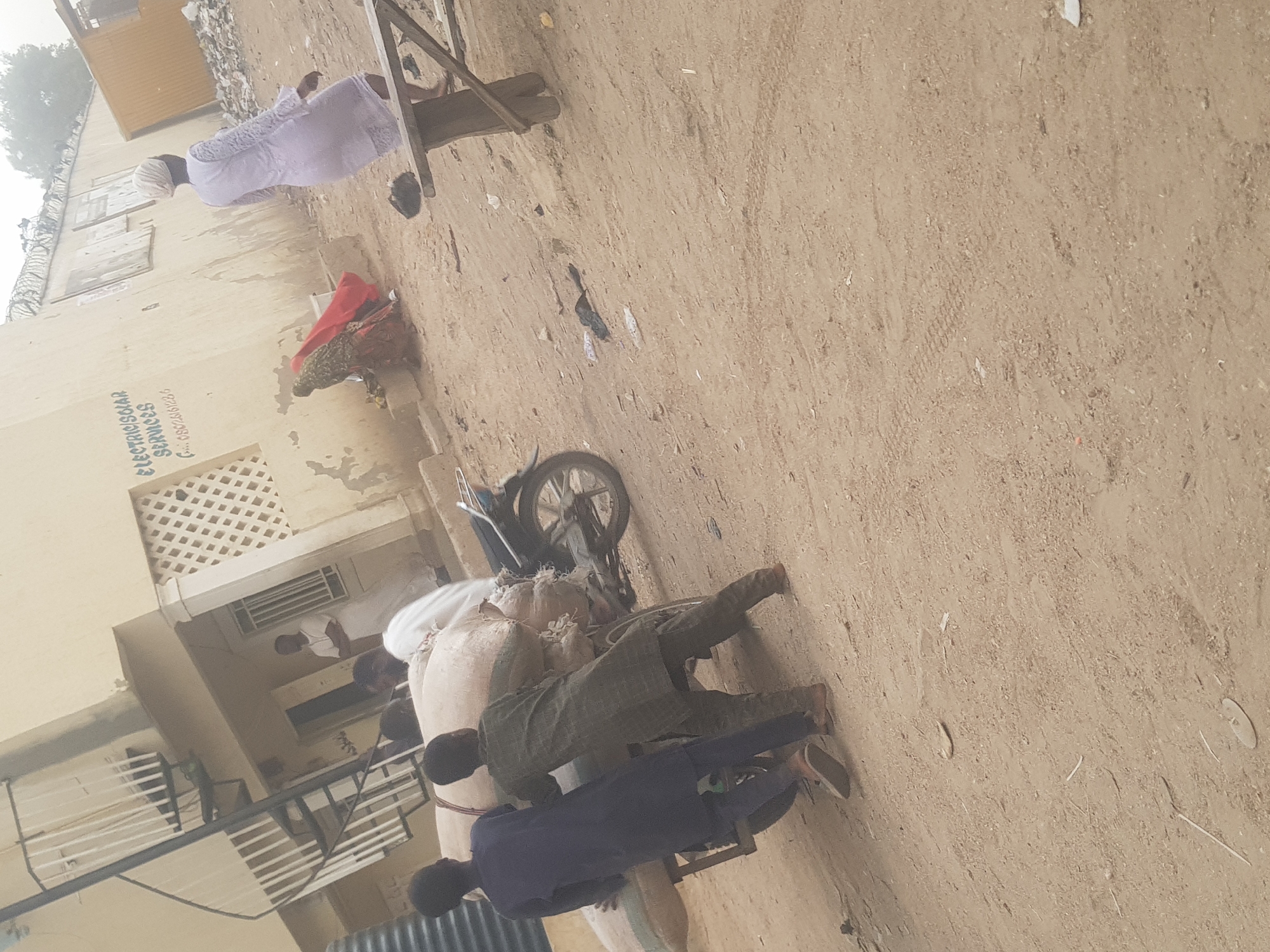 The scene is an outdoor area in Dabawa, Nigeria. The ground is a dirt surface. Several people are present. One person is wearing a long white garment. Two people are seated on a wooden bench. Another person is wearing a red piece of clothing. Further along there is a person wearing a white shirt. Two children wearing a blue and a gray shirt sit on top of bags being transported on a bicycle. The building in the background has a sign that reads "Electrosolar Services".