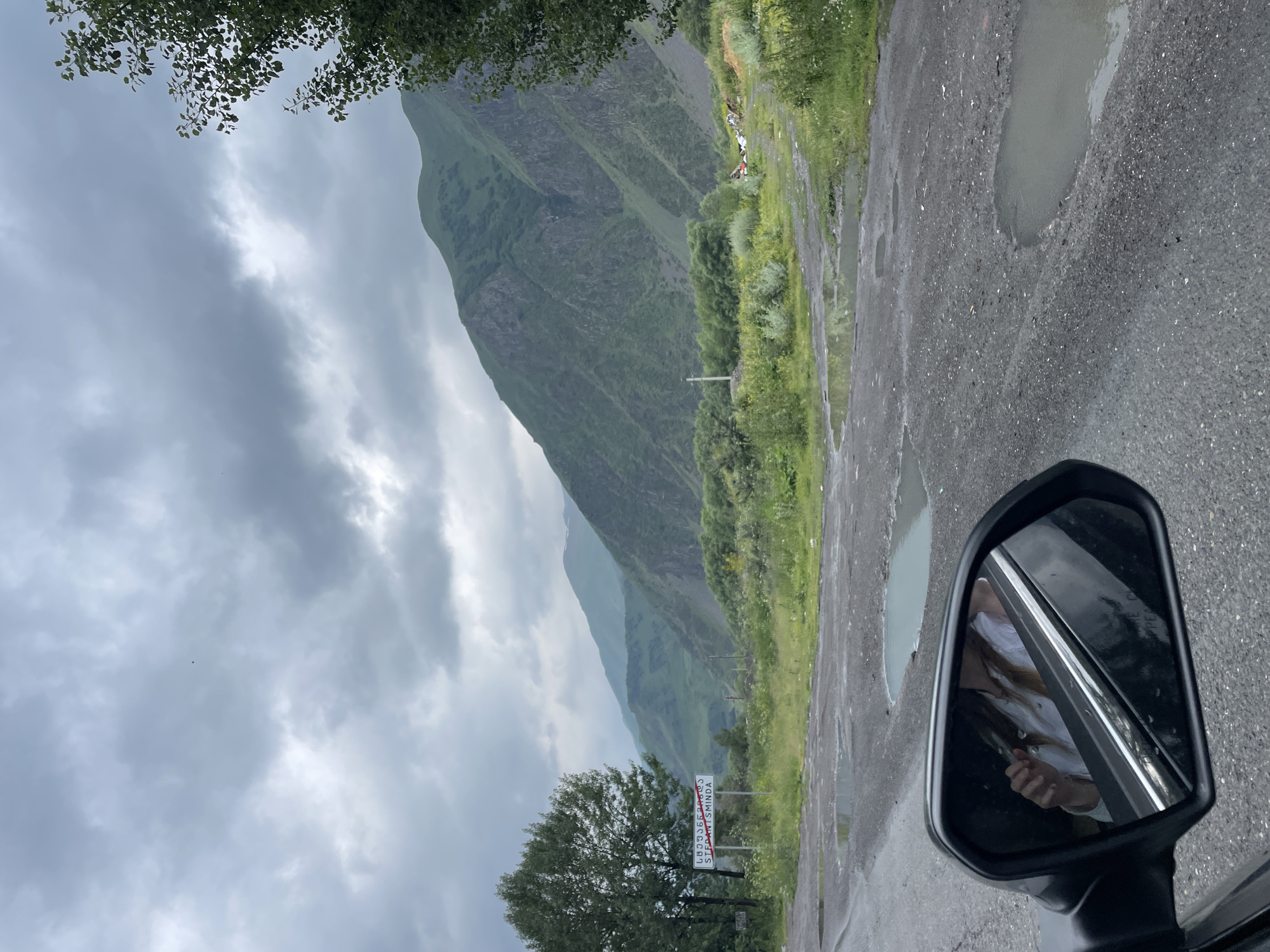 The image shows a view of a road with potholes and puddles in Stepantsminda, Georgia. In the background, there are green mountains under a cloudy sky. A signpost with Georgian script and "STEPHANTSMINDA" written below it is visible on the left side of the road, next to a tree. A side-view mirror of a car is in the foreground, reflecting a person inside the car.