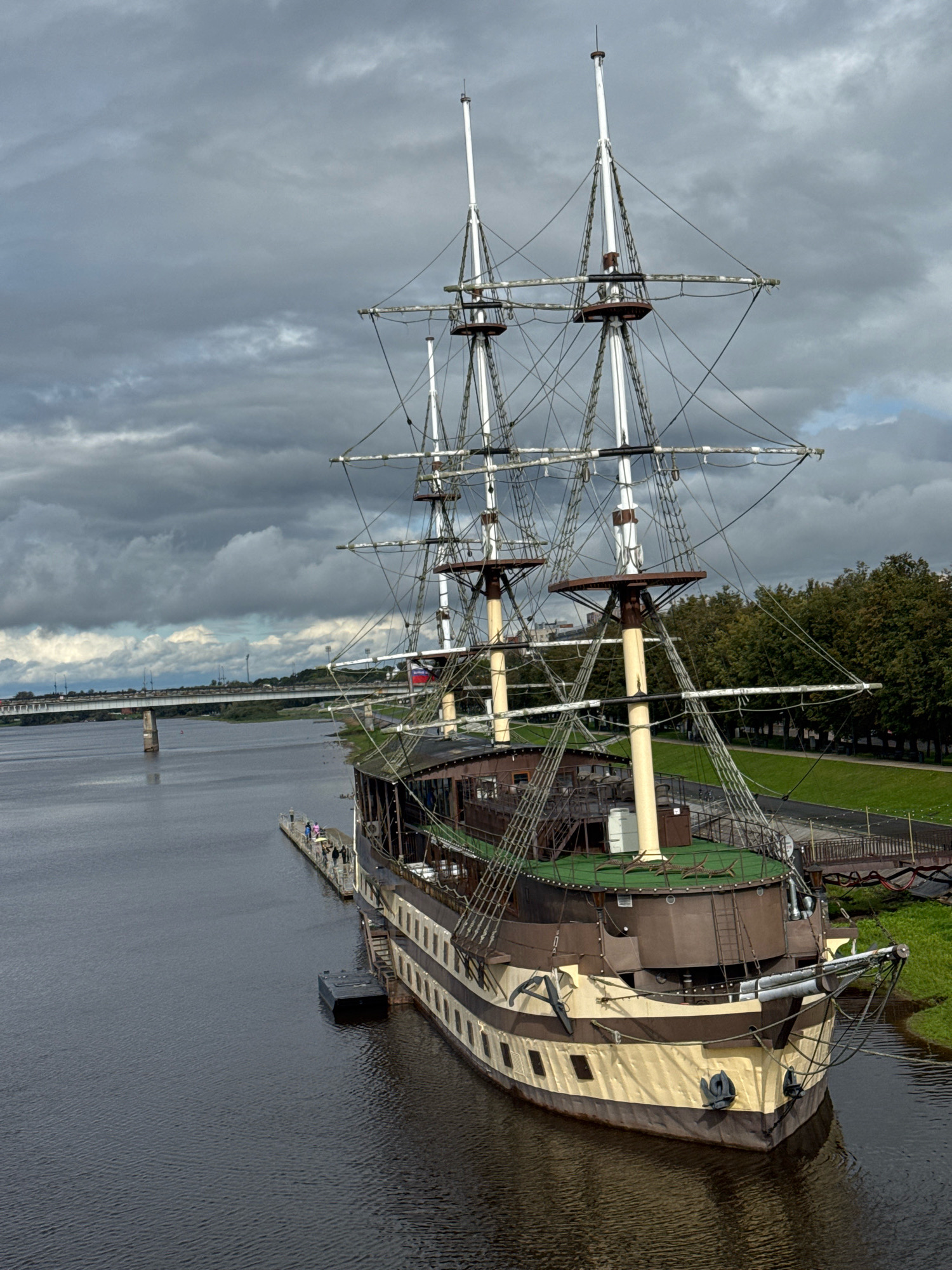 The image shows a historical sailing ship docked on the Volkhov River in Veliky Novgorod, Russia. The ship, named "Krutoy", is a replica of a 17th-century Russian frigate. It is a large, wooden vessel with multiple masts and intricate rigging. The ship is painted in brown and cream colors. Several people are visible on the deck of the ship and on the adjacent pier.
The Volkhov River is wide and calm, with dark, rippling water. On the far bank of the river, a long bridge with multiple arches spans the water, connecting the city to the opposite side. Trees with lush green foliage line the riverbank behind the ship, suggesting a park or recreational area. The sky is overcast with dramatic, grey clouds, indicating a cloudy day. The scene appears to be a tourist attraction, with the ship serving as a museum or restaurant.