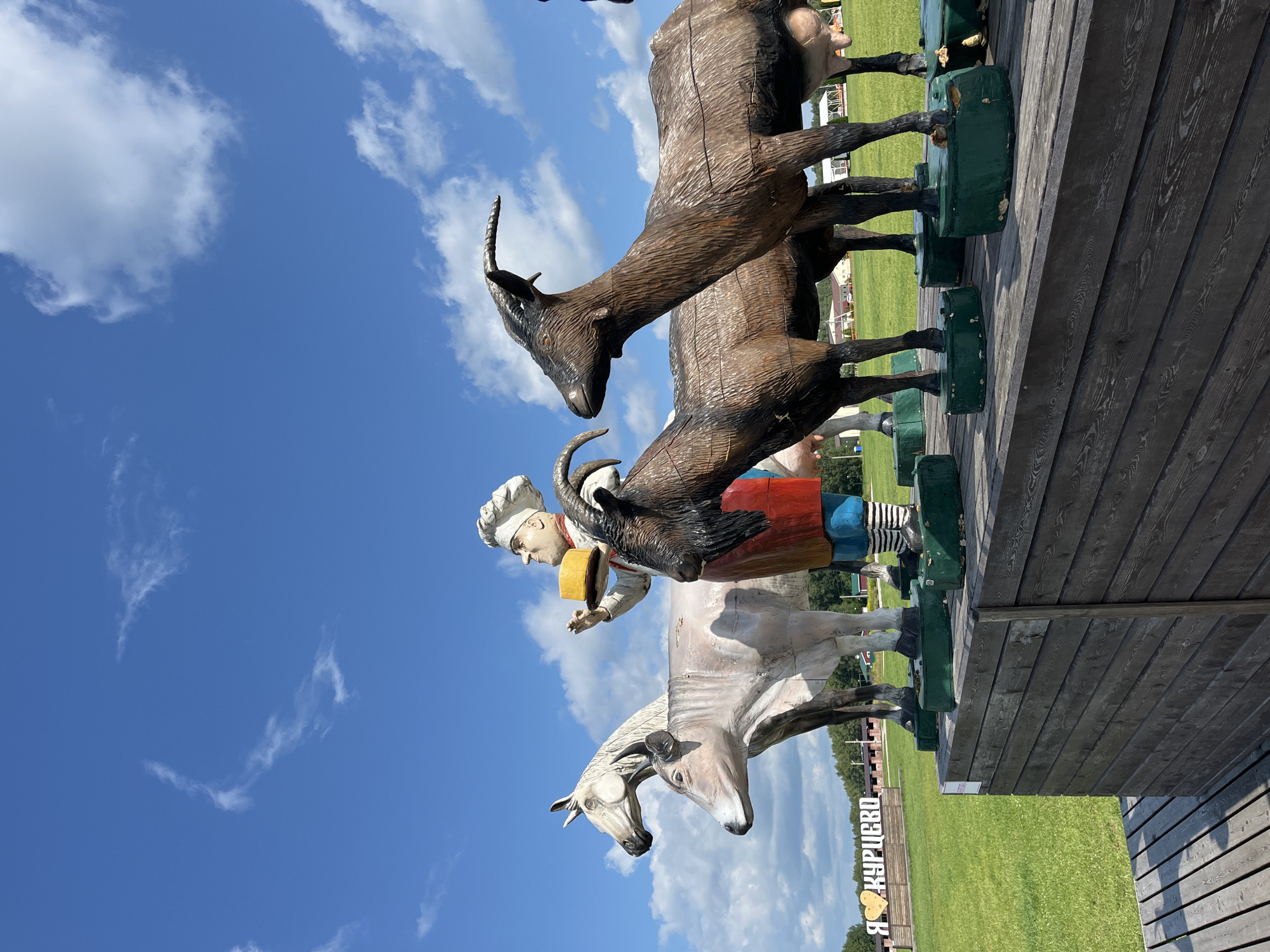 The image depicts a wooden sculpture of a chef holding a round of cheese, standing in front of a white horse and a cow. Two goats stand to the right of the chef. The sculptures are mounted on a wooden platform, which is part of an outdoor exhibition. In the background, a green field stretches out, with trees and buildings visible in the distance. A large white sign with the Cyrillic text "Я КУРИЦЕВО" (Ya Kuritsevo) and a heart symbol is also visible in the background. The sky is blue with scattered white clouds. This scene is located in Akhlebaikha (Kovriginskii S/S), Russia, and is likely a tourist attraction.