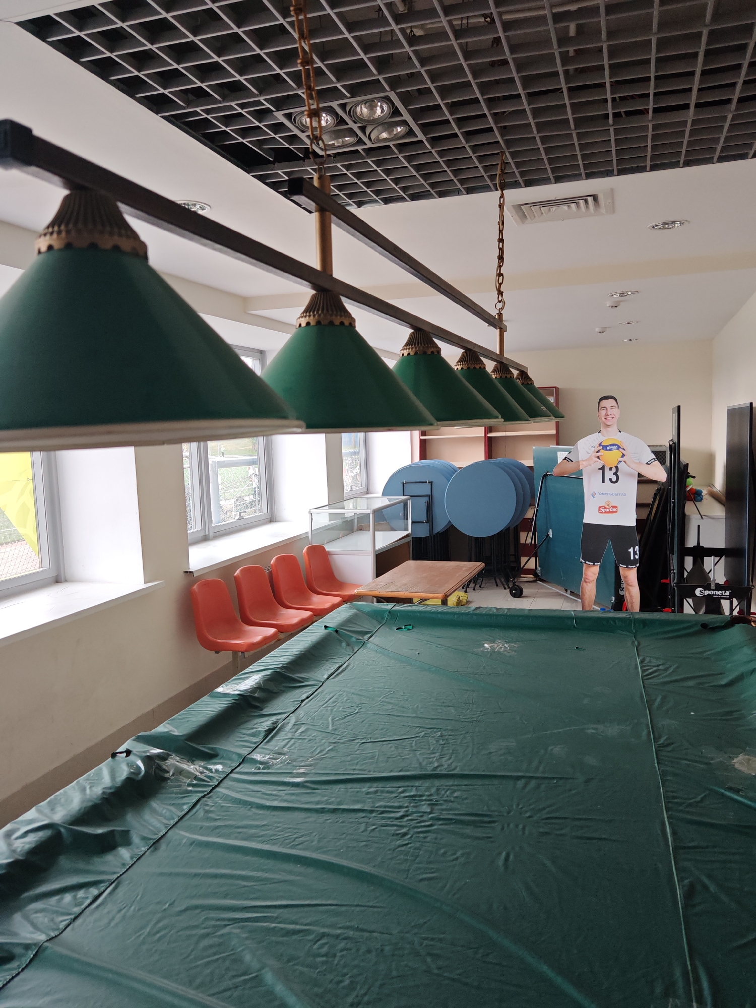 A lifesized cardboard cutout of a male volleyball player, wearing a white t-shirt with "13" on it and black shorts, stands on the right side of the frame, holding a volleyball. In front of the cutout is a large, green-covered pool table, likely for billiards or snooker. Several orange chairs are lined up along the left wall, which features large windows. The room has a high, coffered ceiling with pendant lights hanging over the pool table. In the background, stacked round tables and shelving units are visible. The overall impression is that of a recreational area or a hall in Homyel, Belarus, possibly a sports club or community center.