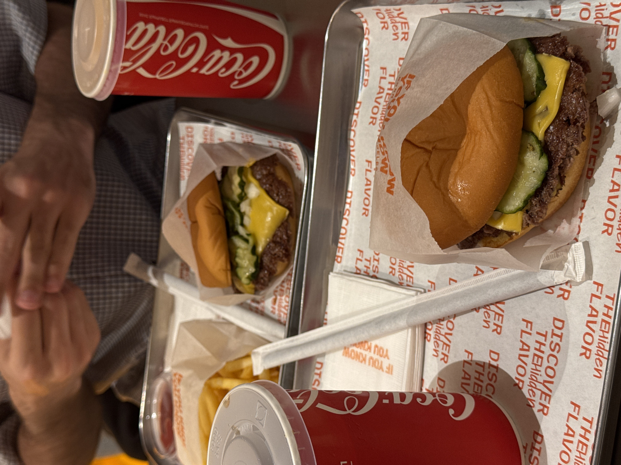 The image shows a person's hands at a table with two burgers, fries, and two Coca-Cola drinks on trays. The trays are covered with paper printed with the text "DISCOVER THE Hidden FLAVOR". The burgers have cheese and pickles. No specific location in Yerevan, Armenia, can be determined from the image.